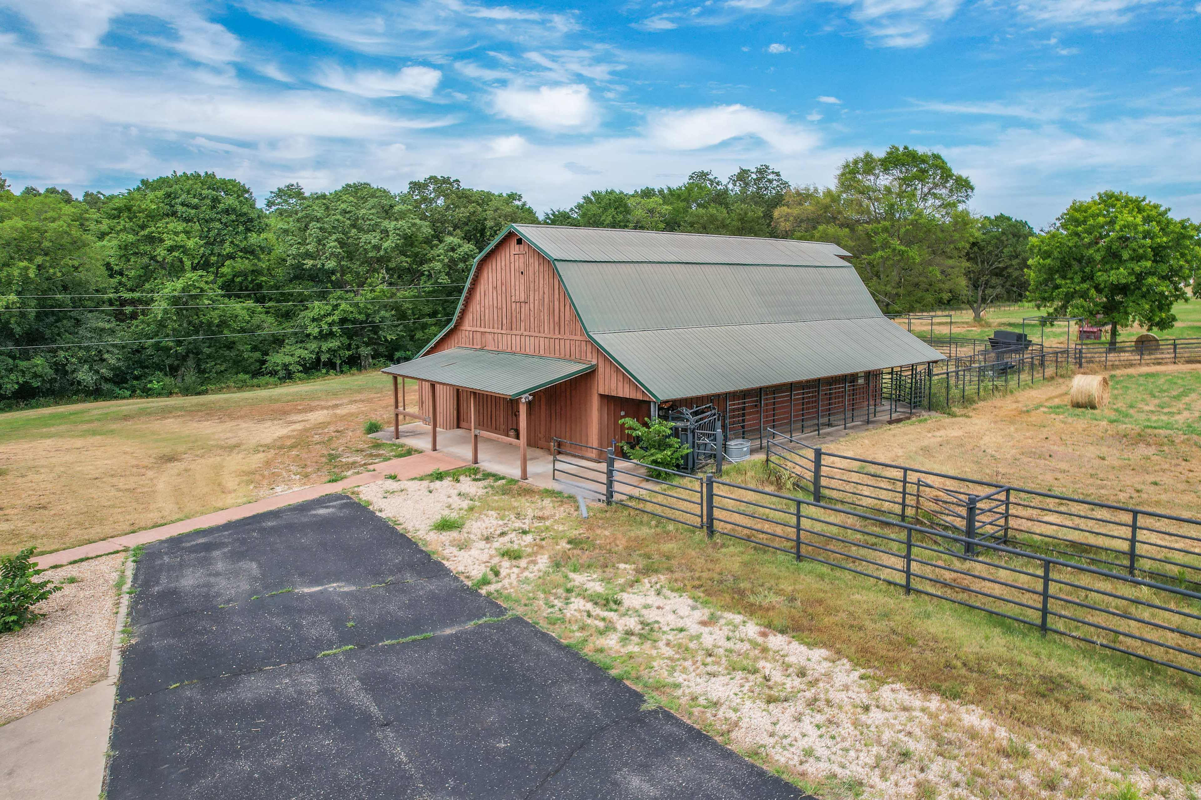 A large barn with a green metal roof situated on a grassy field, surrounded by trees and a fenced area.