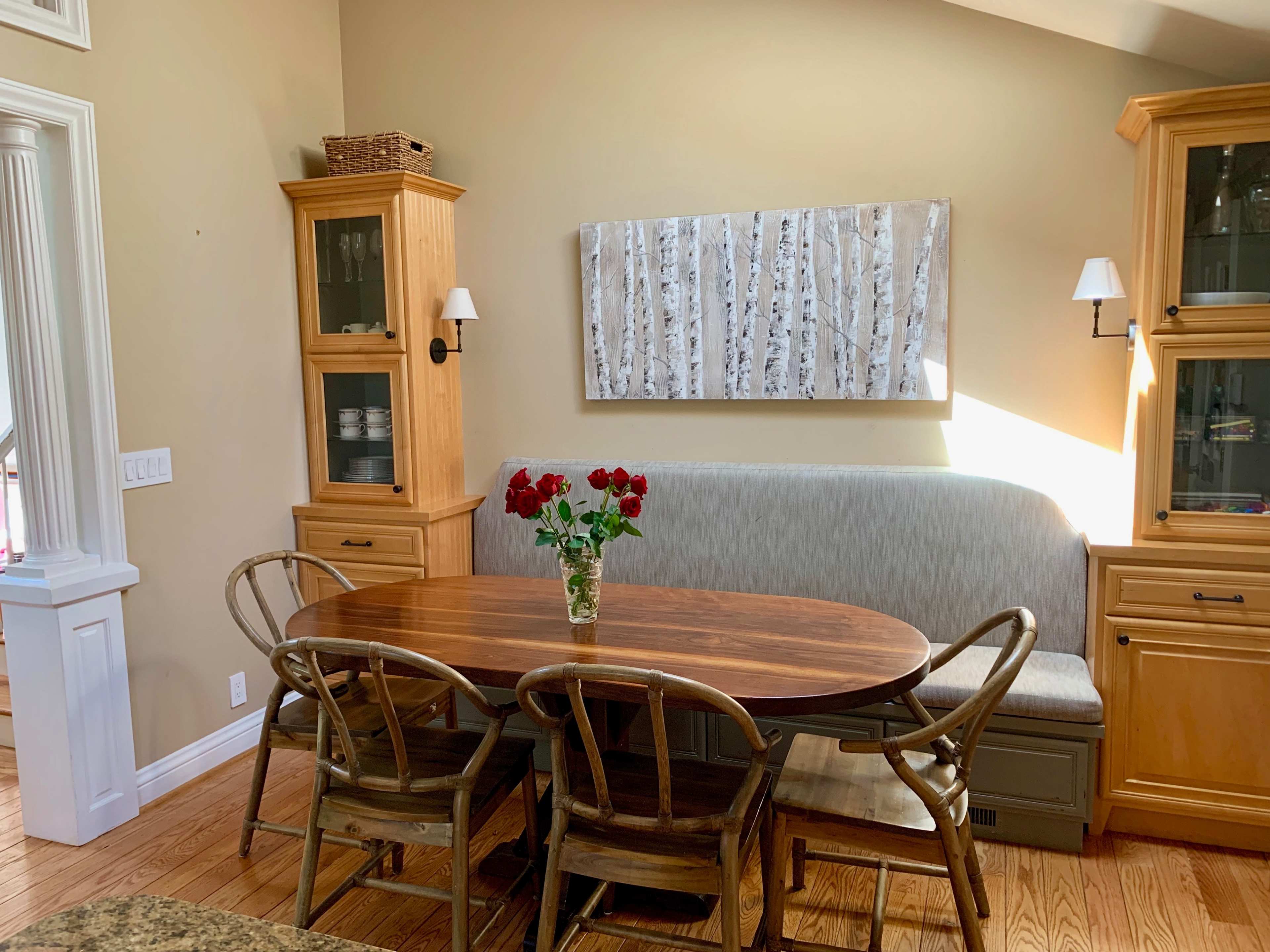 A wooden dining table surrounded by chairs is set in a brightly lit room, featuring a floral centerpiece and a wall-mounted artwork above a cushioned bench.