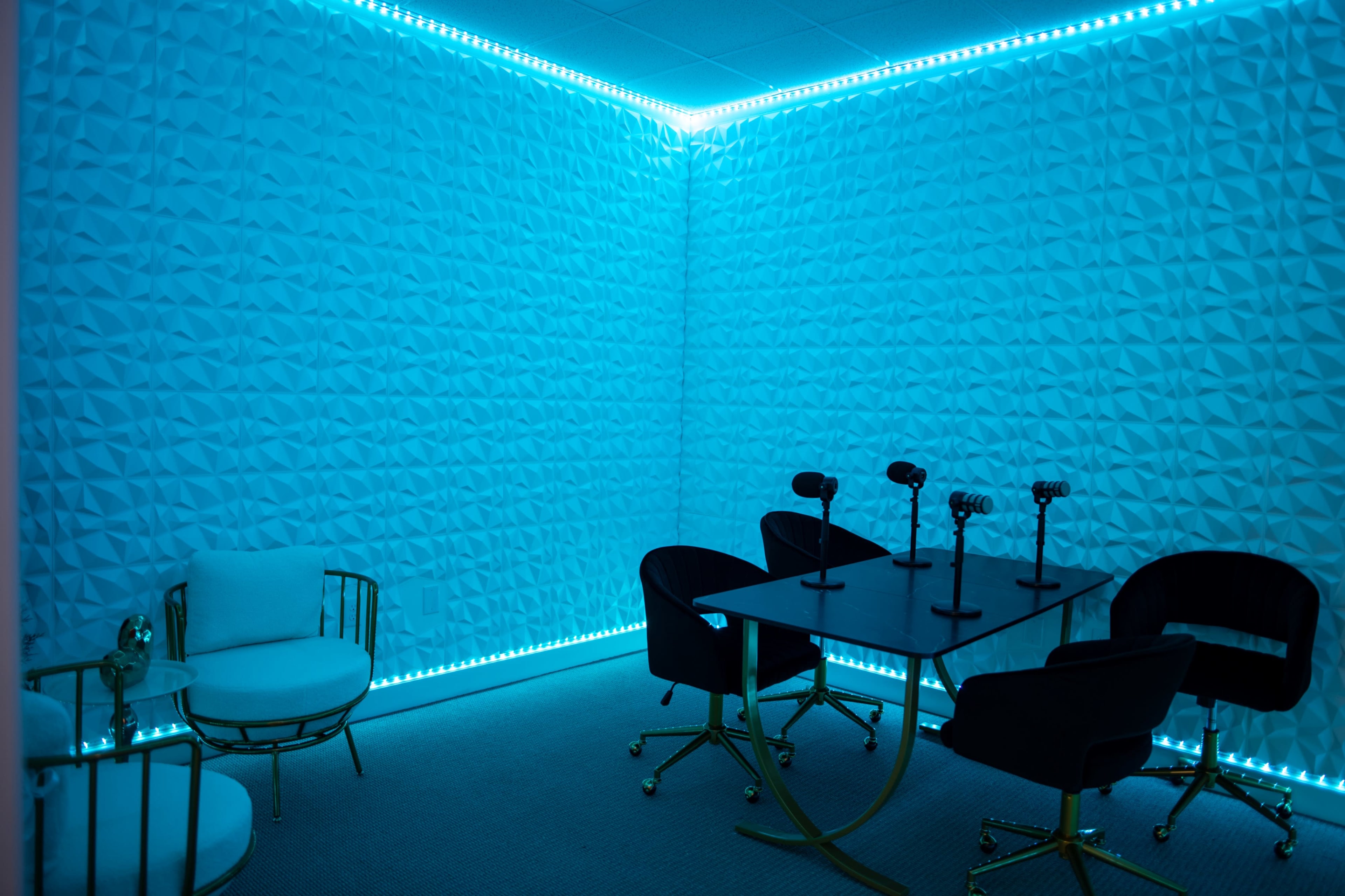 The image shows a brightly lit room with textured blue walls, a modern black table surrounded by four chairs, and soft seating in one corner.