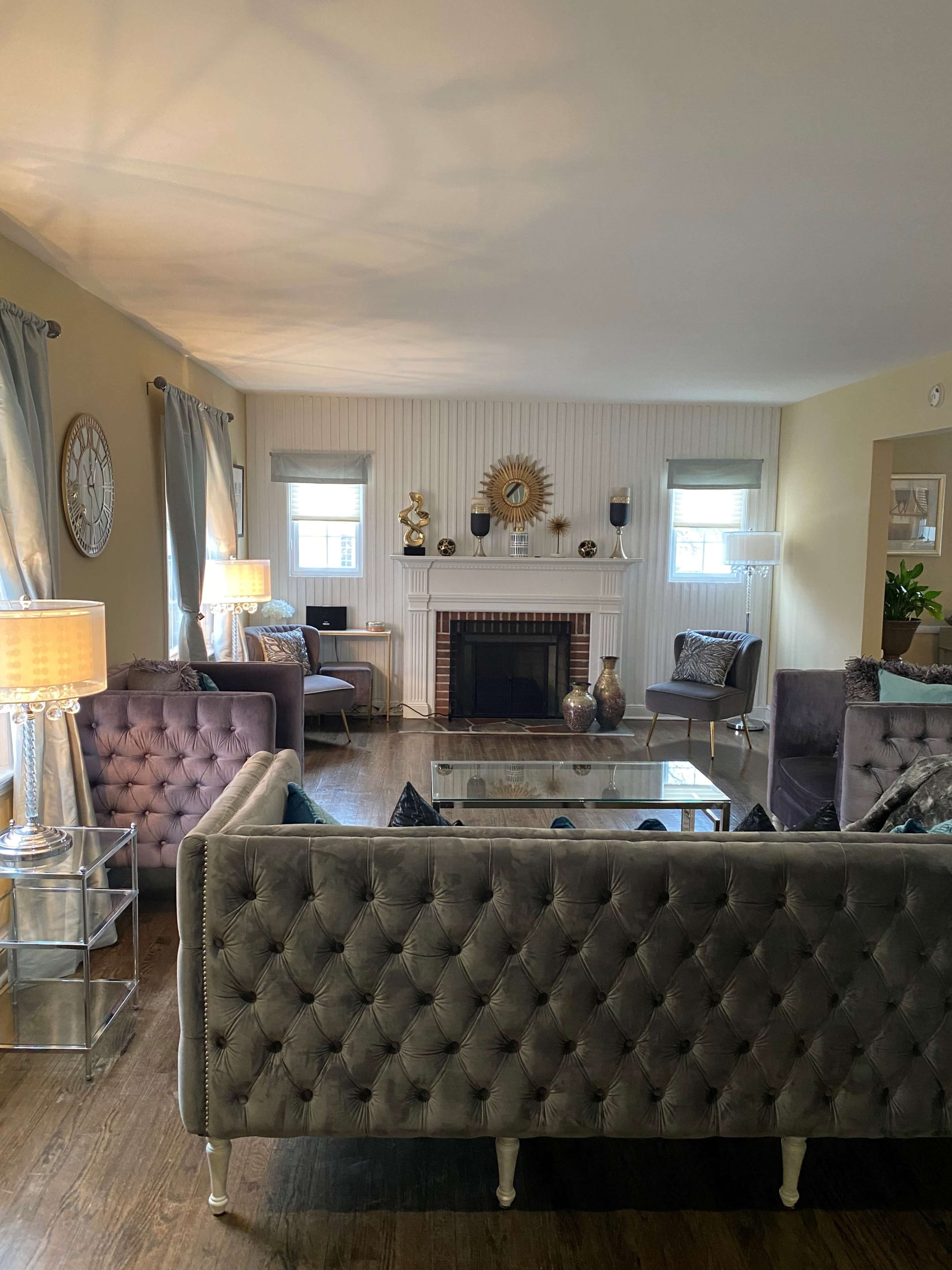A well-furnished living room featuring a gray tufted sofa, a glass coffee table, a fireplace, and several decorative items arranged on the mantel.