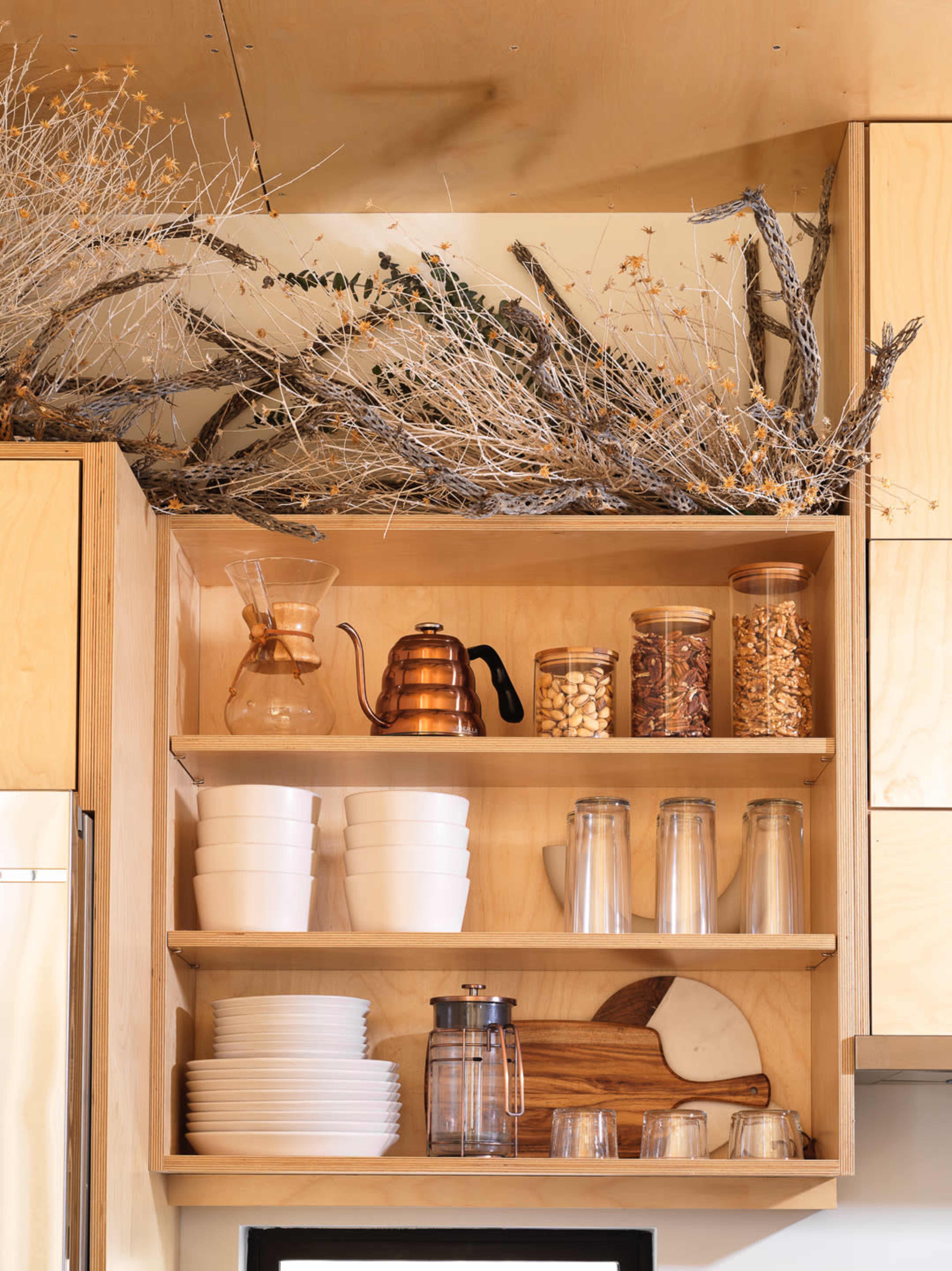 A wooden kitchen shelf displays stacked white plates, glassware, jars of nuts, and a copper kettle, topped with dry branches and foliage.