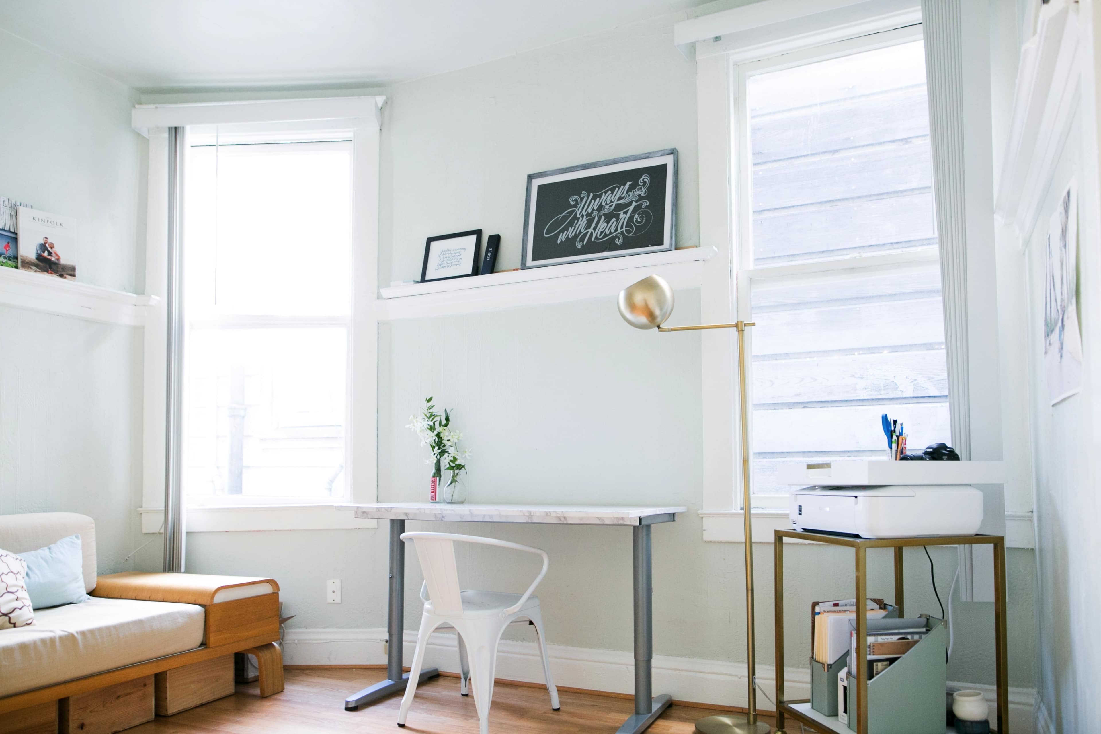 The room features a minimalist workspace with a wooden desk, a white chair, and a printer beside a small plant, illuminated by natural light from two windows.