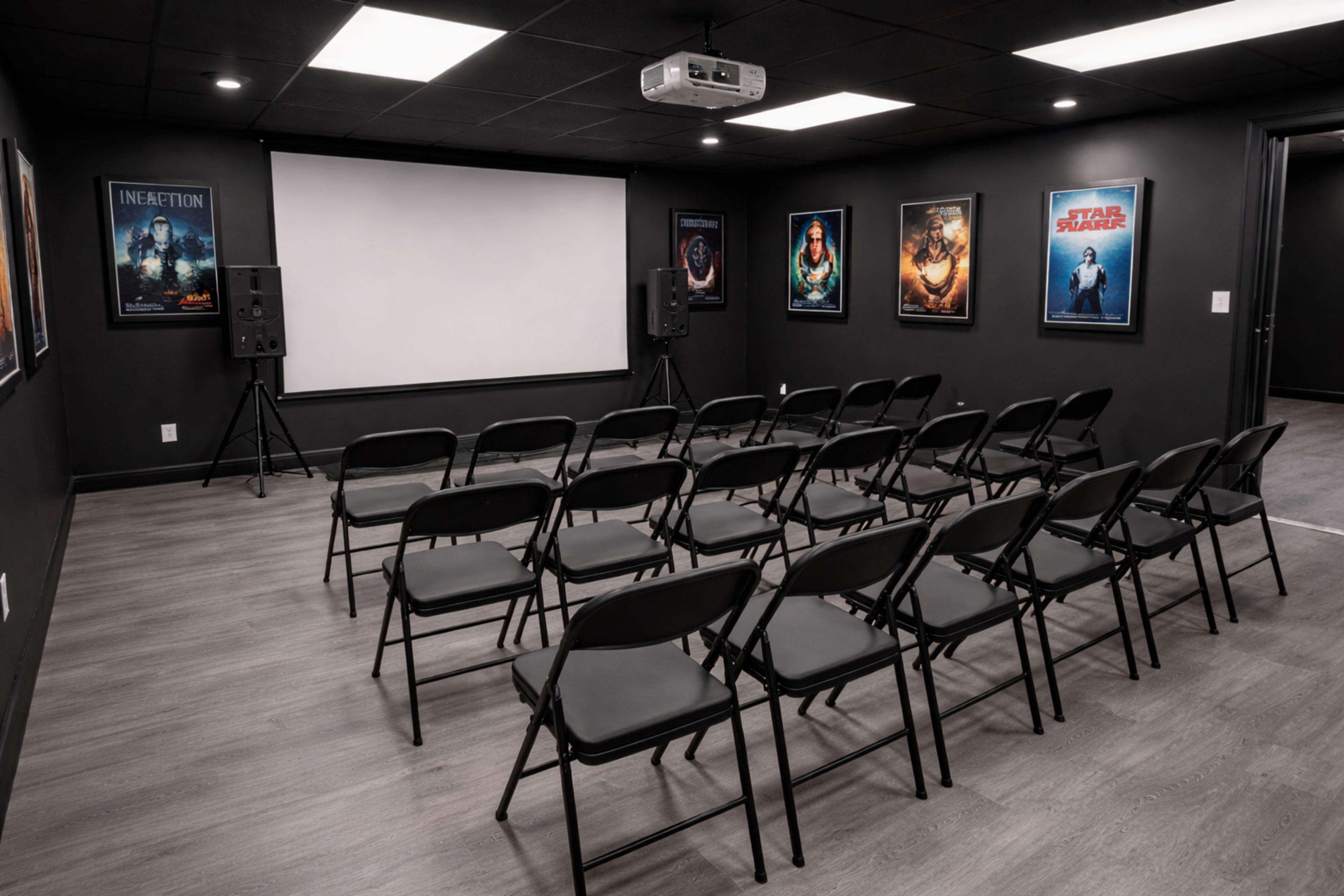 The image shows a dark room arranged with rows of black folding chairs facing a large projection screen, flanked by movie posters on the walls.