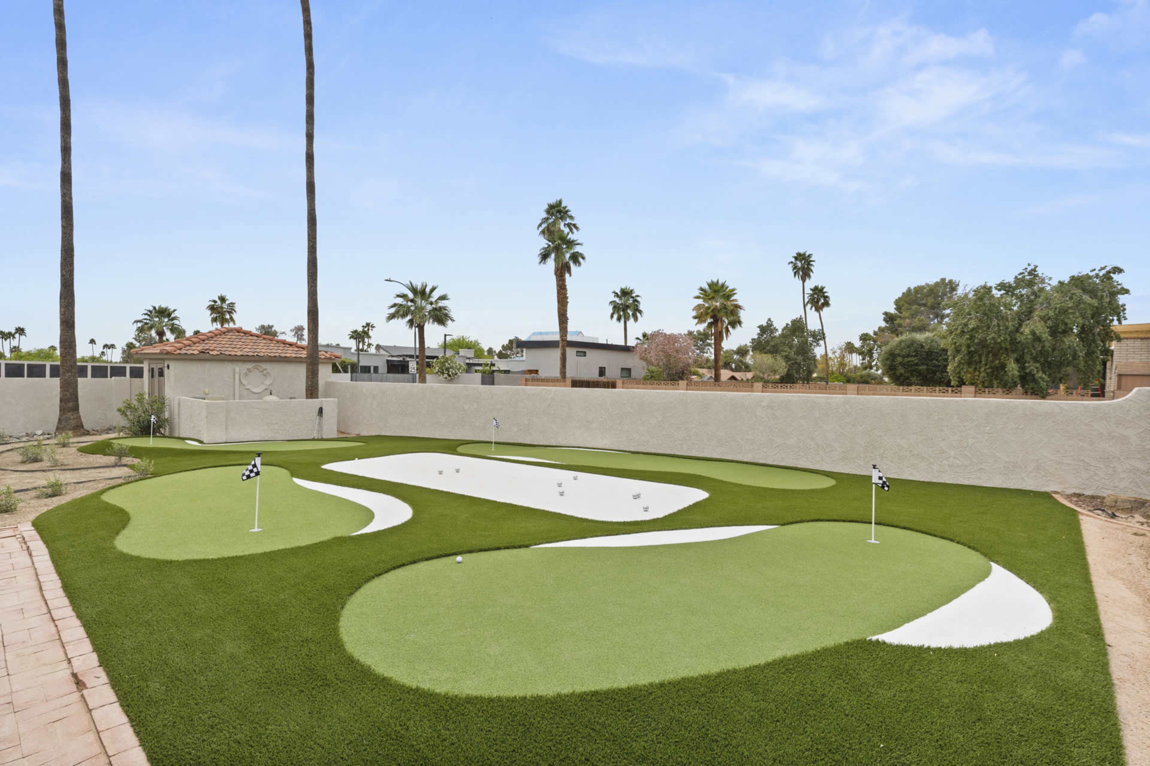 The image shows a residential backyard featuring two putting green areas with cup holes, surrounded by artificial grass and framed by palm trees.