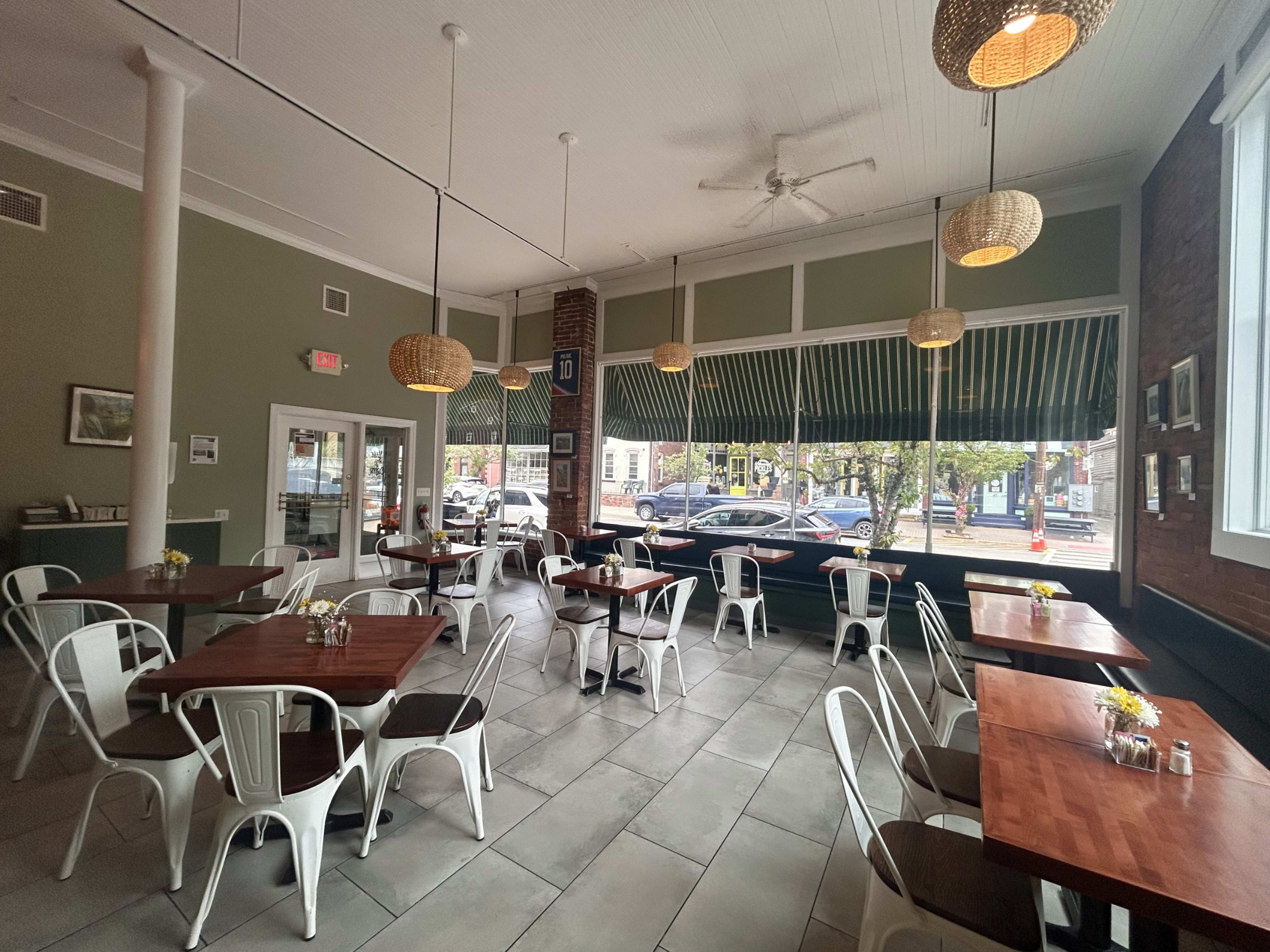 The interior of a spacious, contemporary café features wooden tables and metal chairs, with large windows overlooking a sidewalk and street.