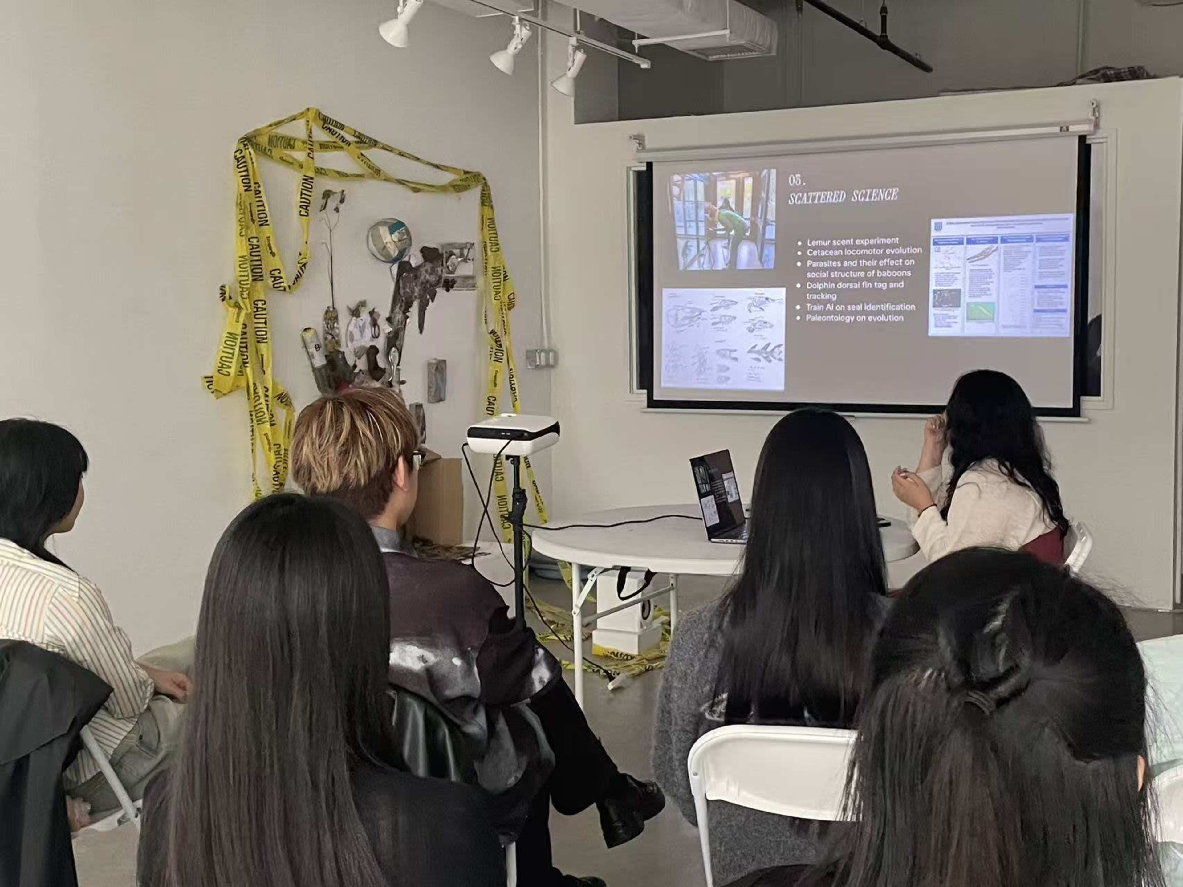 A small group of people is seated in front of a projector screen displaying information in a room decorated with caution tape and art pieces.