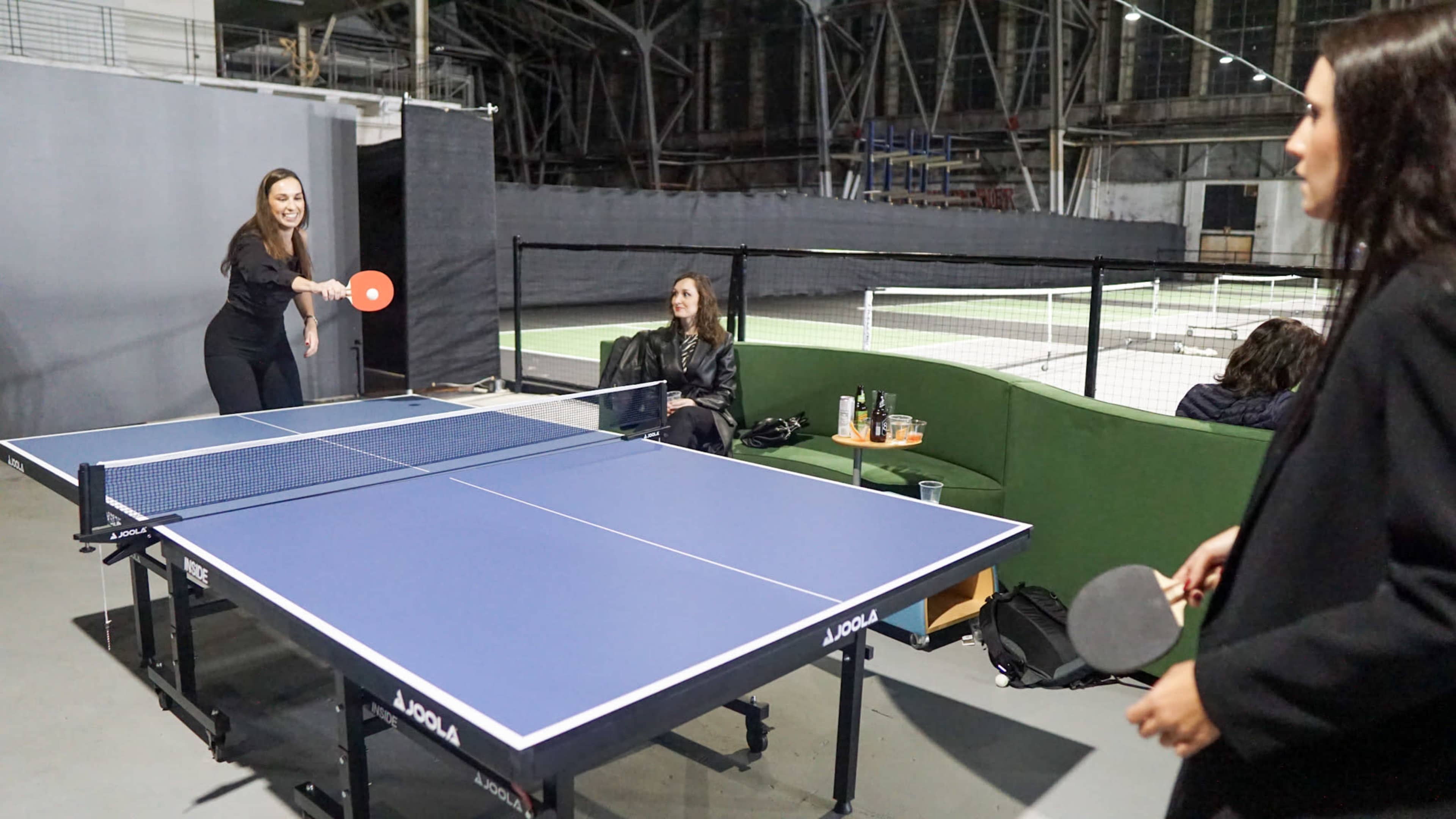 Two women play table tennis indoors while another woman observes from a nearby seating area.
