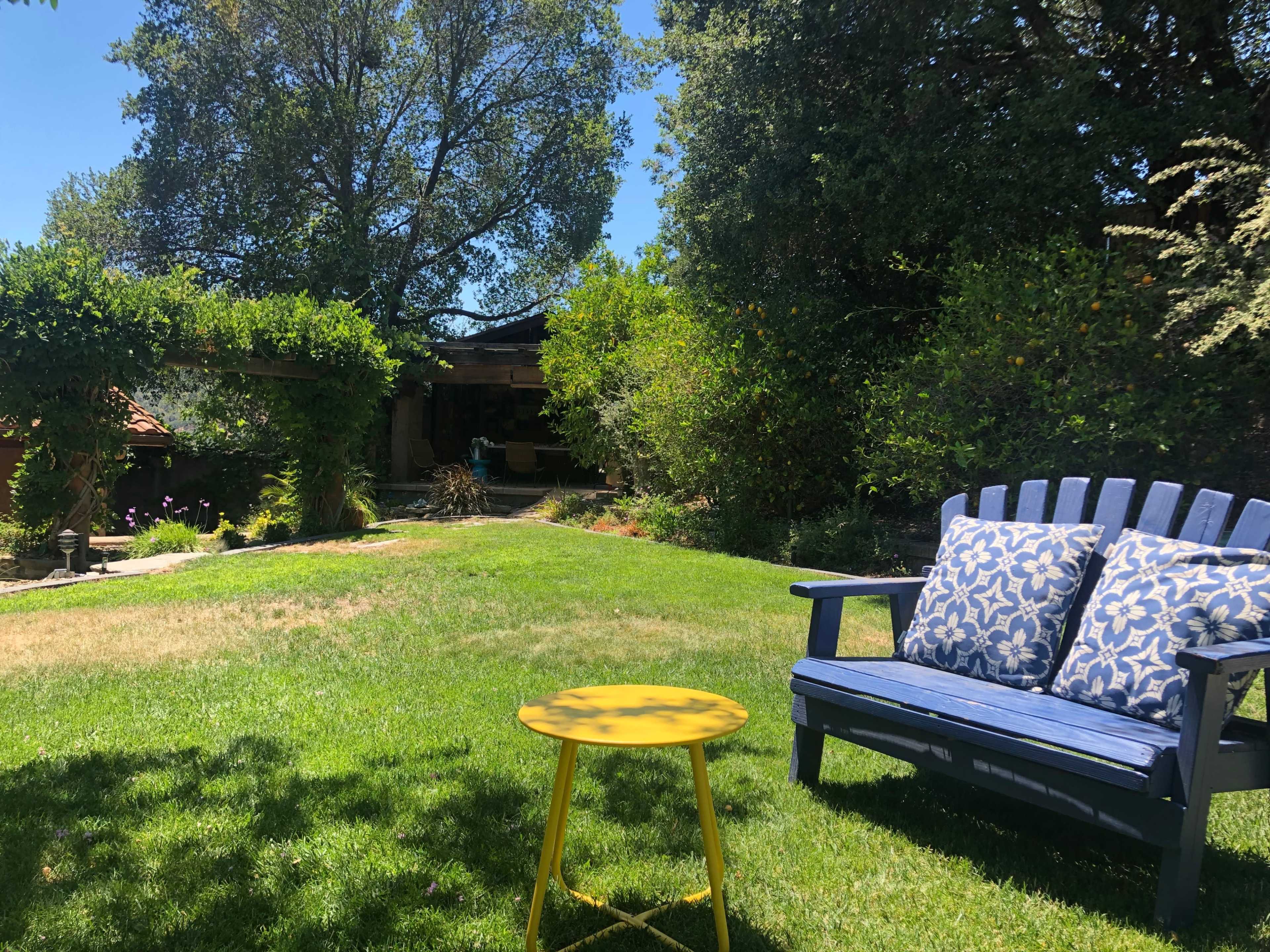 A blue wooden bench with patterned cushions and a yellow side table sits on a grassy lawn surrounded by trees and shrubs.