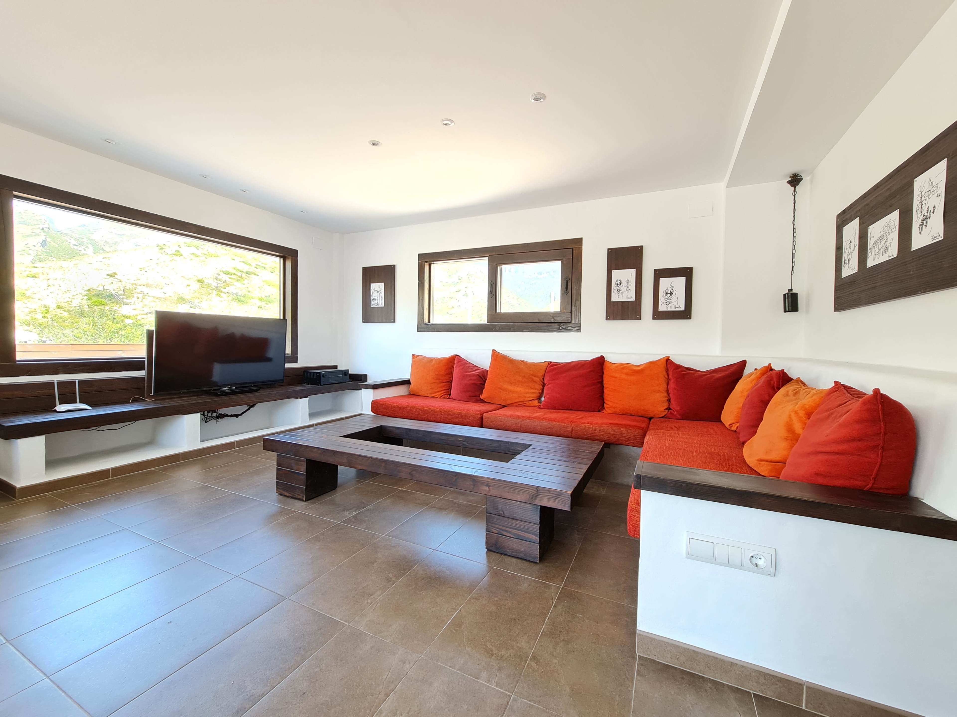 The image shows a modern living room with a large orange sectional sofa, a wooden coffee table, and a TV mounted on the wall, surrounded by large windows letting in natural light.