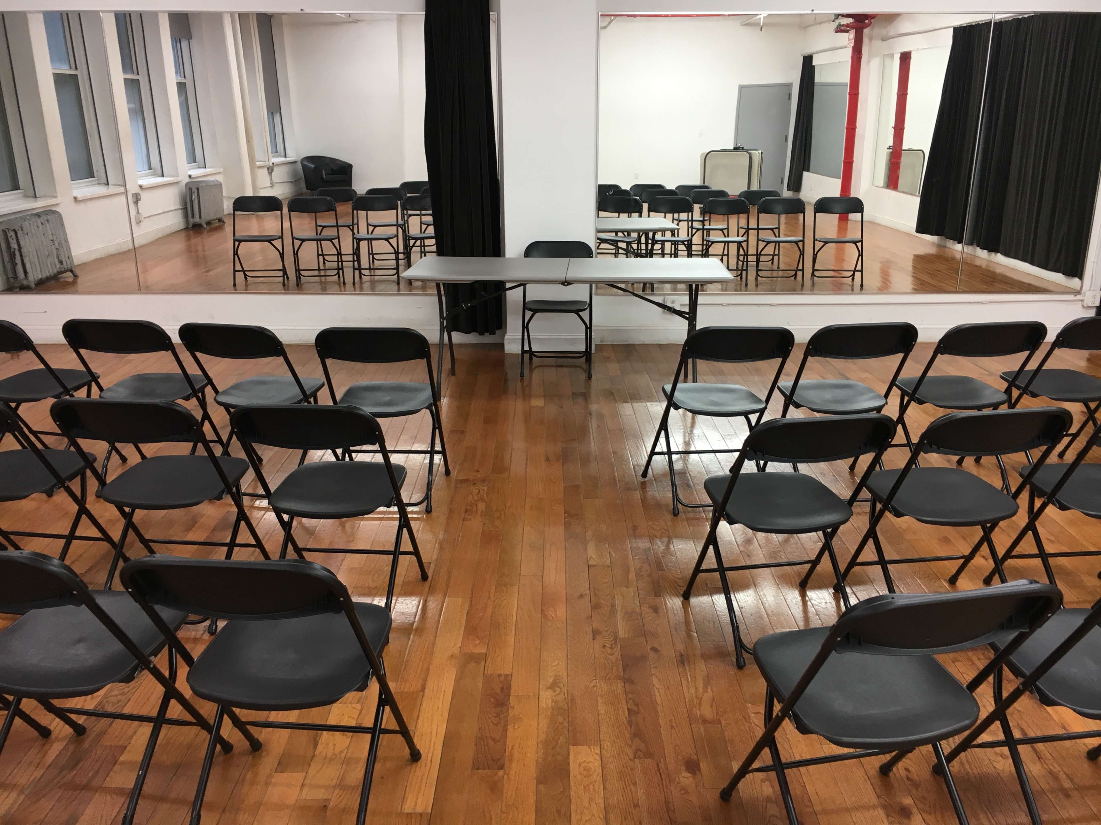 A room with wooden flooring, where black folding chairs are arranged in rows facing a table, with mirrors and additional chairs visible in the background.
