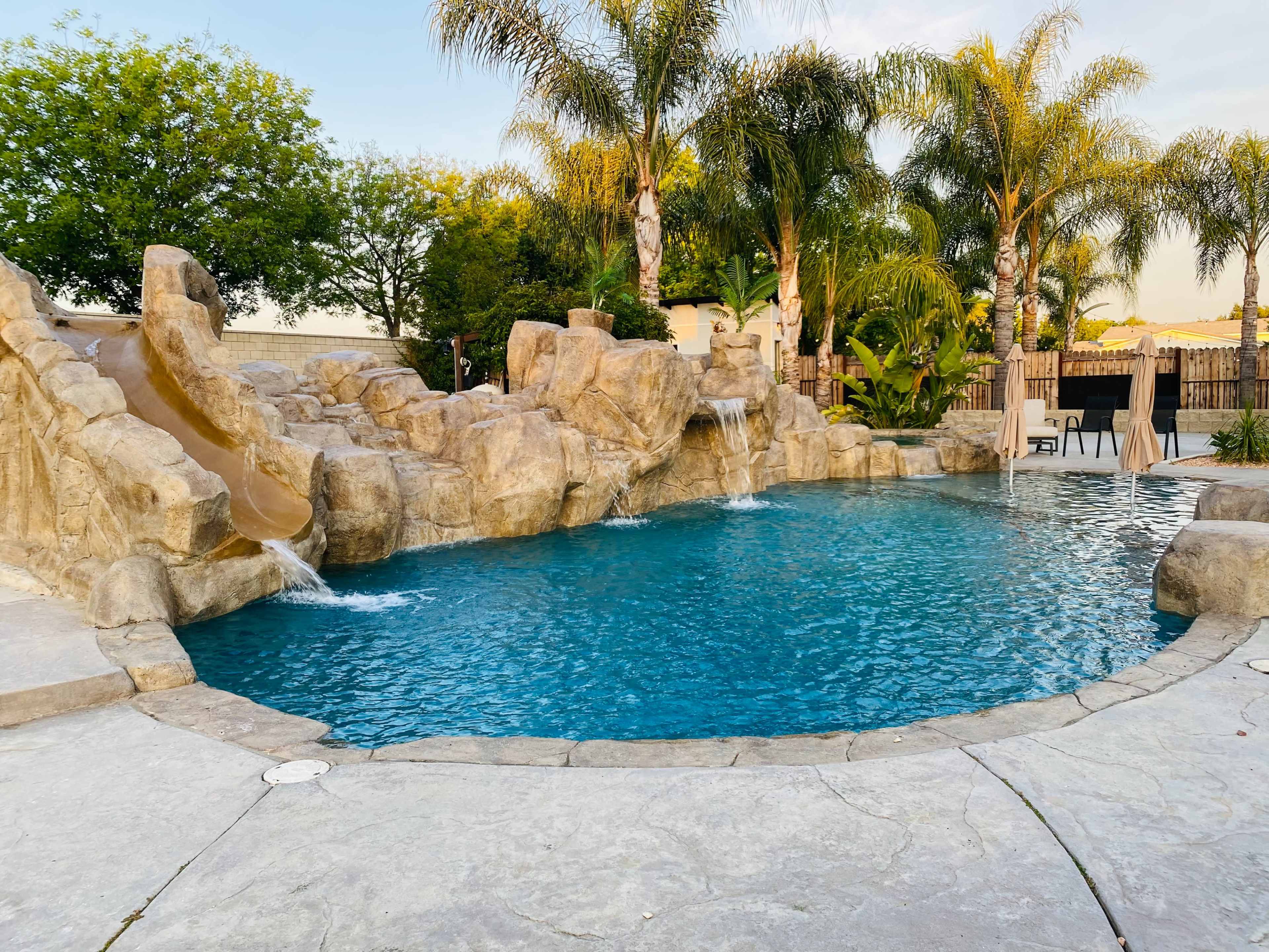 The image shows a swimming pool with a rock-shaped slide, surrounded by palm trees and a landscaped area.