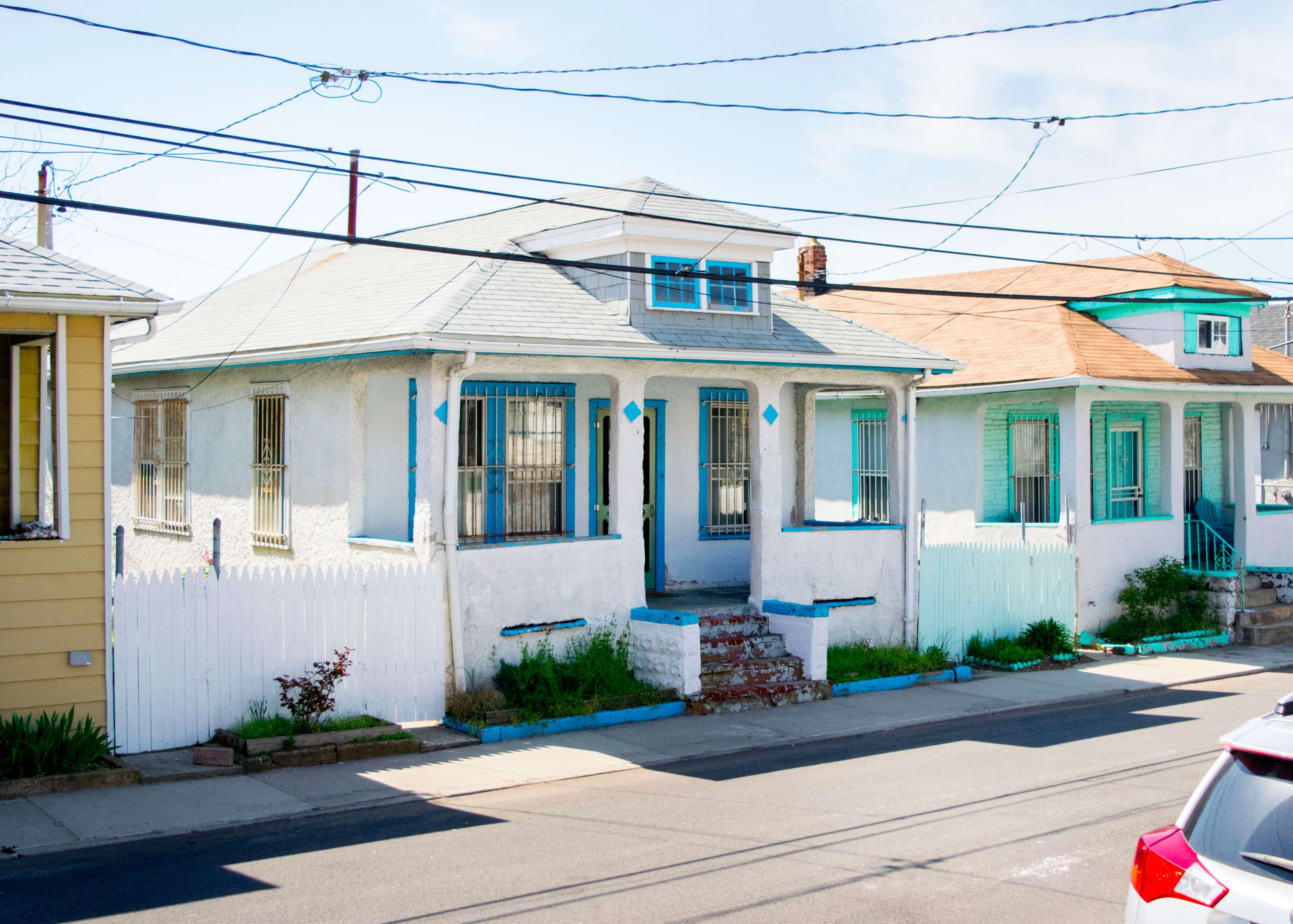 A white stucco house with blue accents sits beside two neighboring houses, all surrounded by grass and a low white picket fence.