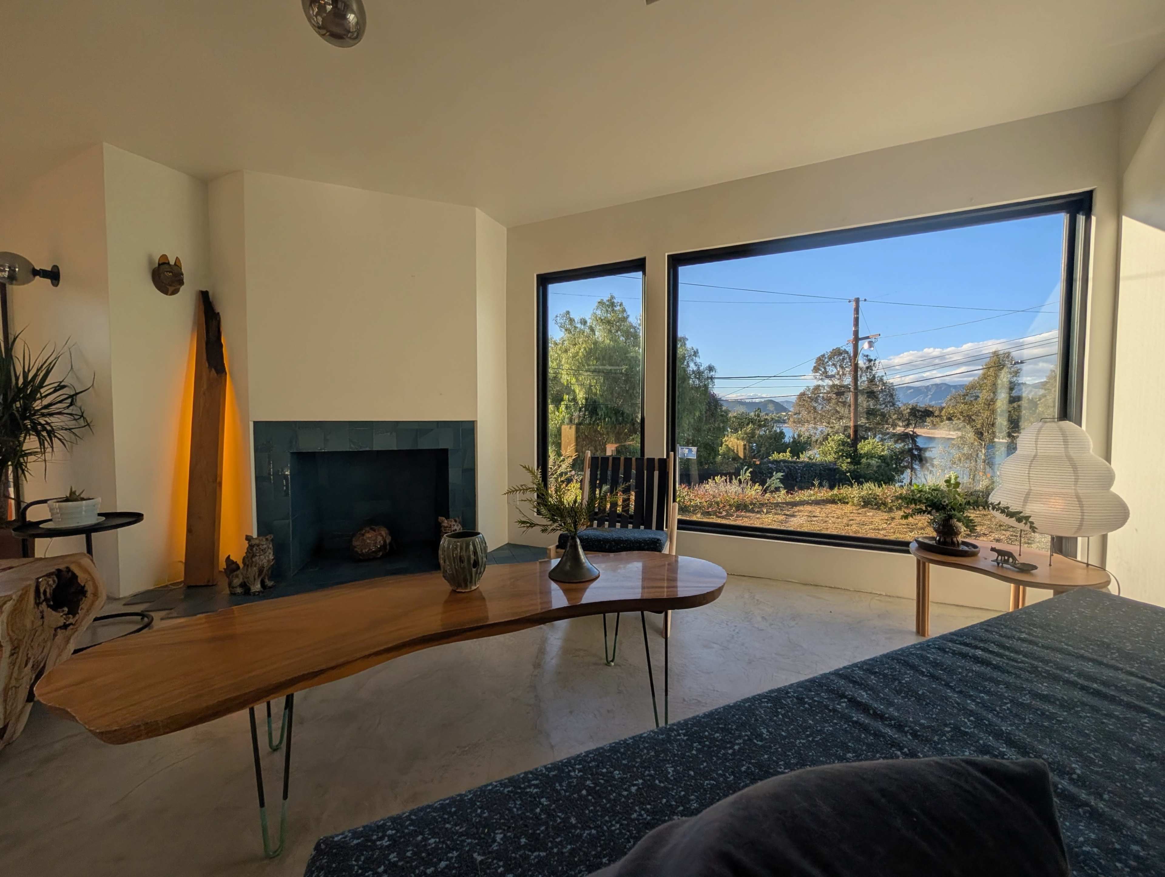 The image shows a modern living room with a large window showcasing a view of greenery and a body of water outside, complemented by a wooden table and minimalist decor.