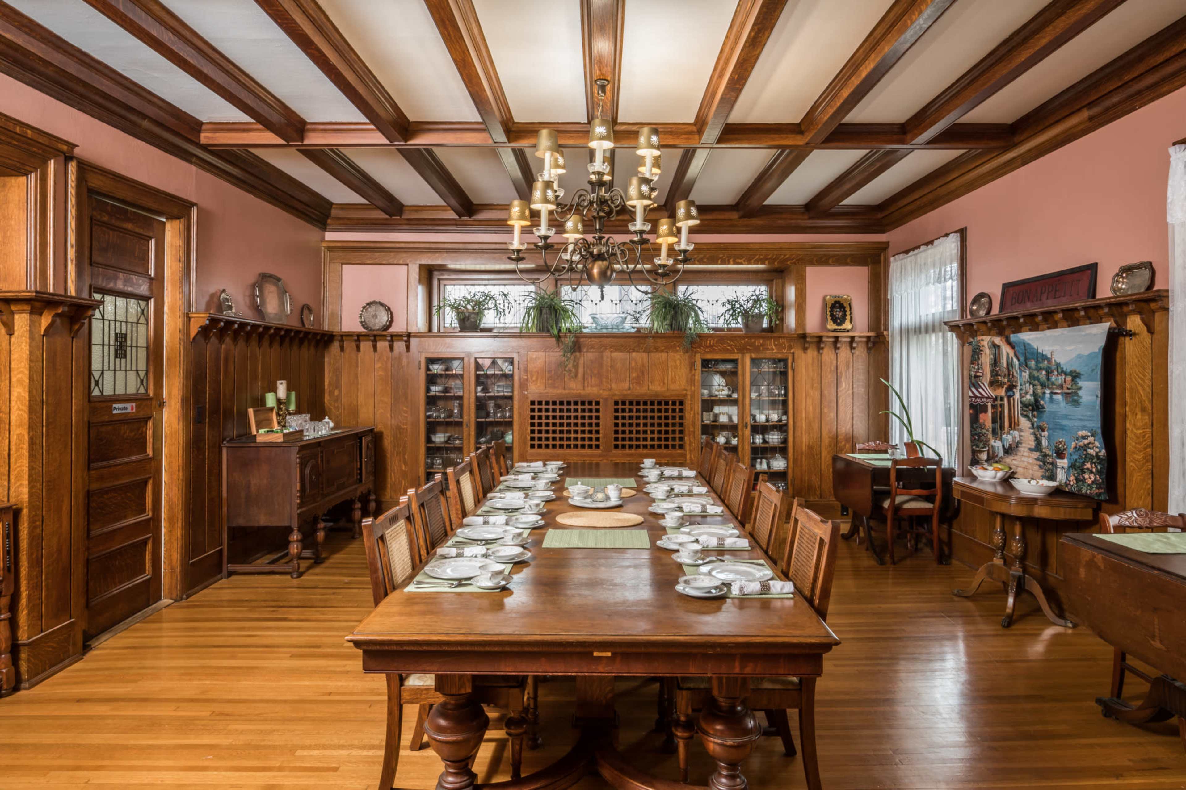 A formal dining room with a long wooden table set for a meal, surrounded by wooden chairs, and featuring chandeliers and decorative woodwork.