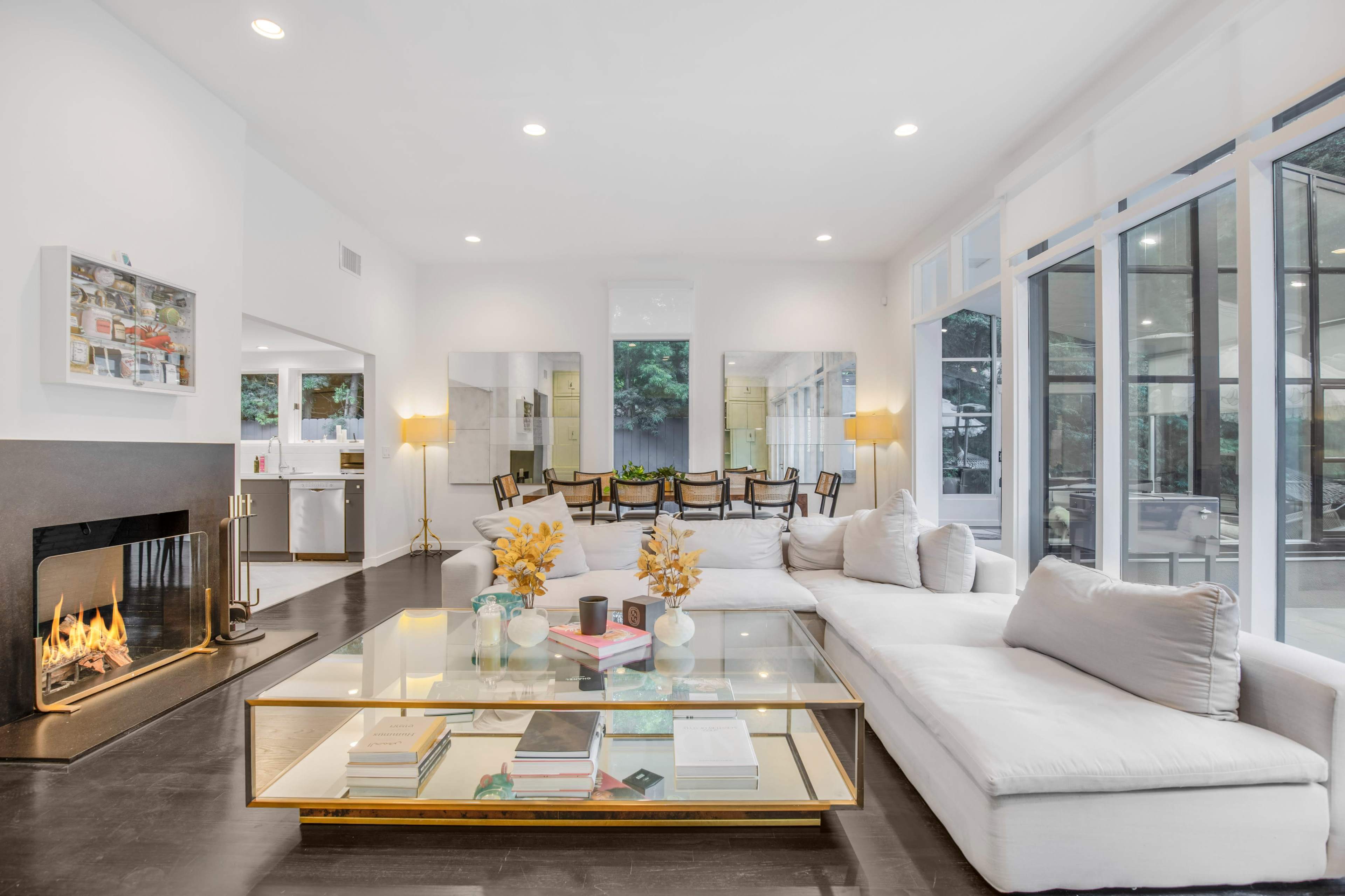 The image shows a modern living room with a large sectional sofa, a glass coffee table, a fireplace, and large windows that let in natural light.
