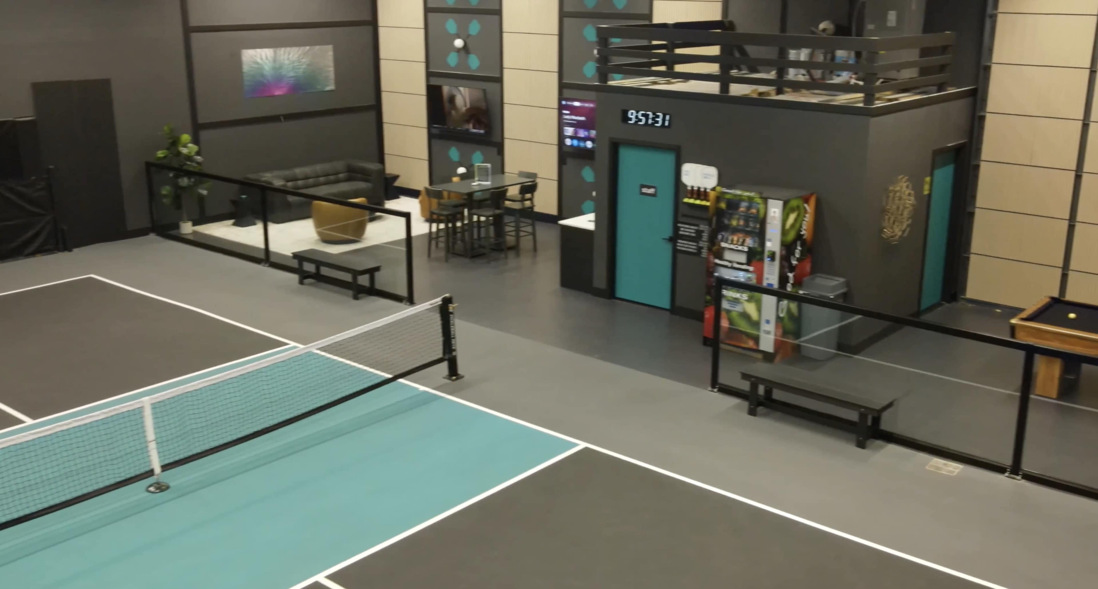 The image shows an indoor sports facility featuring a tennis court with a net, surrounded by seating areas and vending machines.
