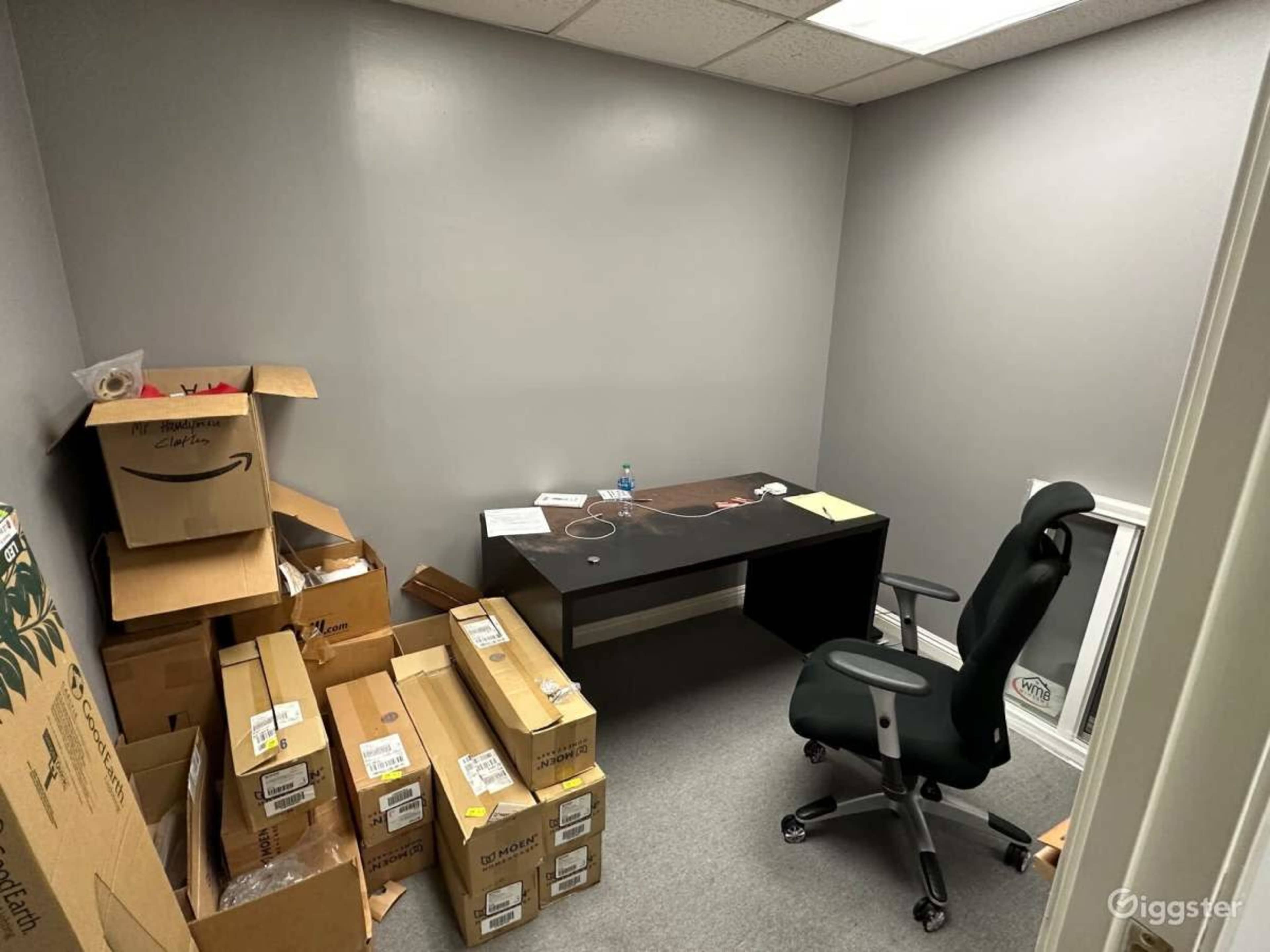 A small office space with a black desk, an ergonomic chair, and several cardboard boxes stacked against the walls.
