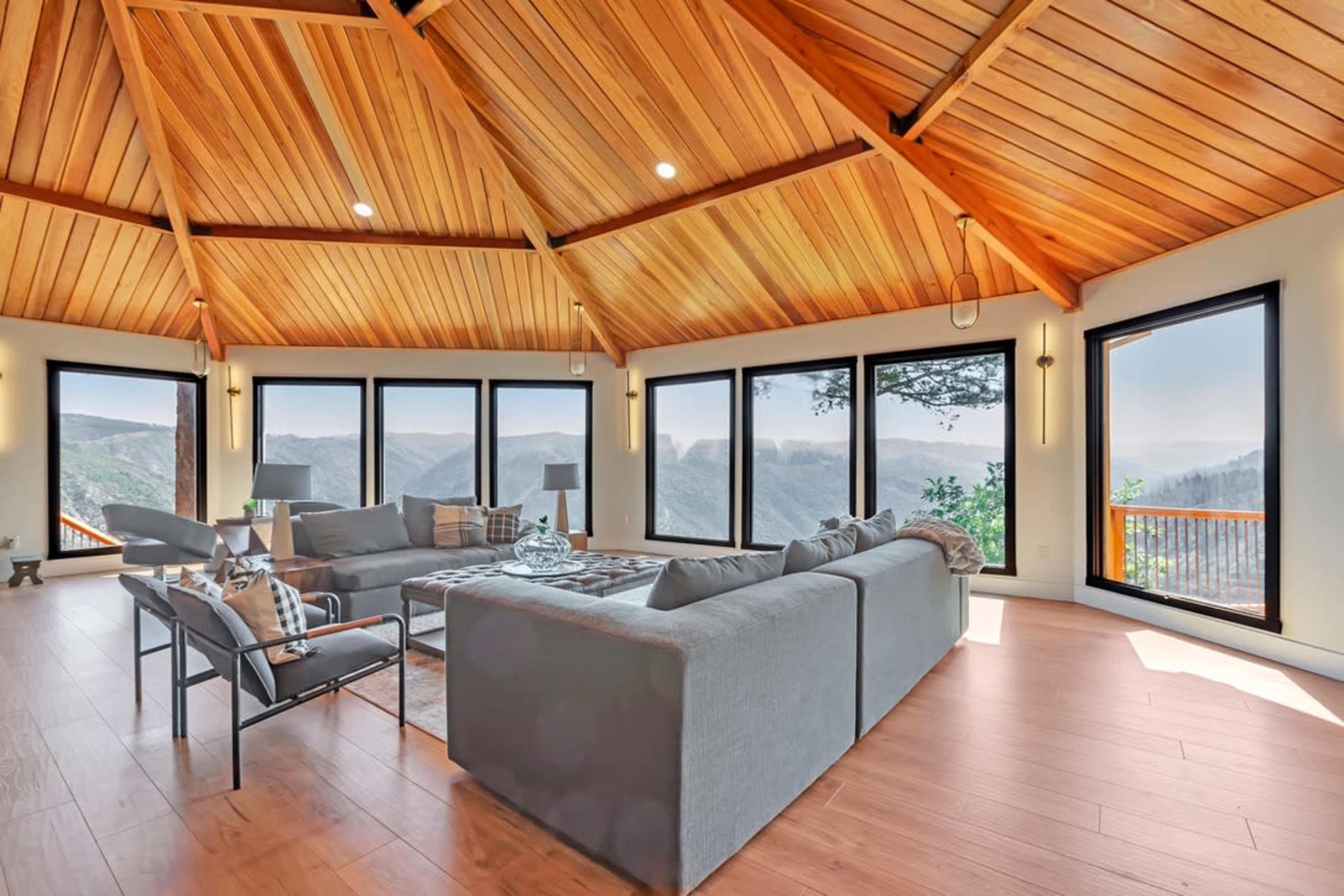 A spacious living room with a high wooden ceiling and large windows overlooking a mountainous landscape.