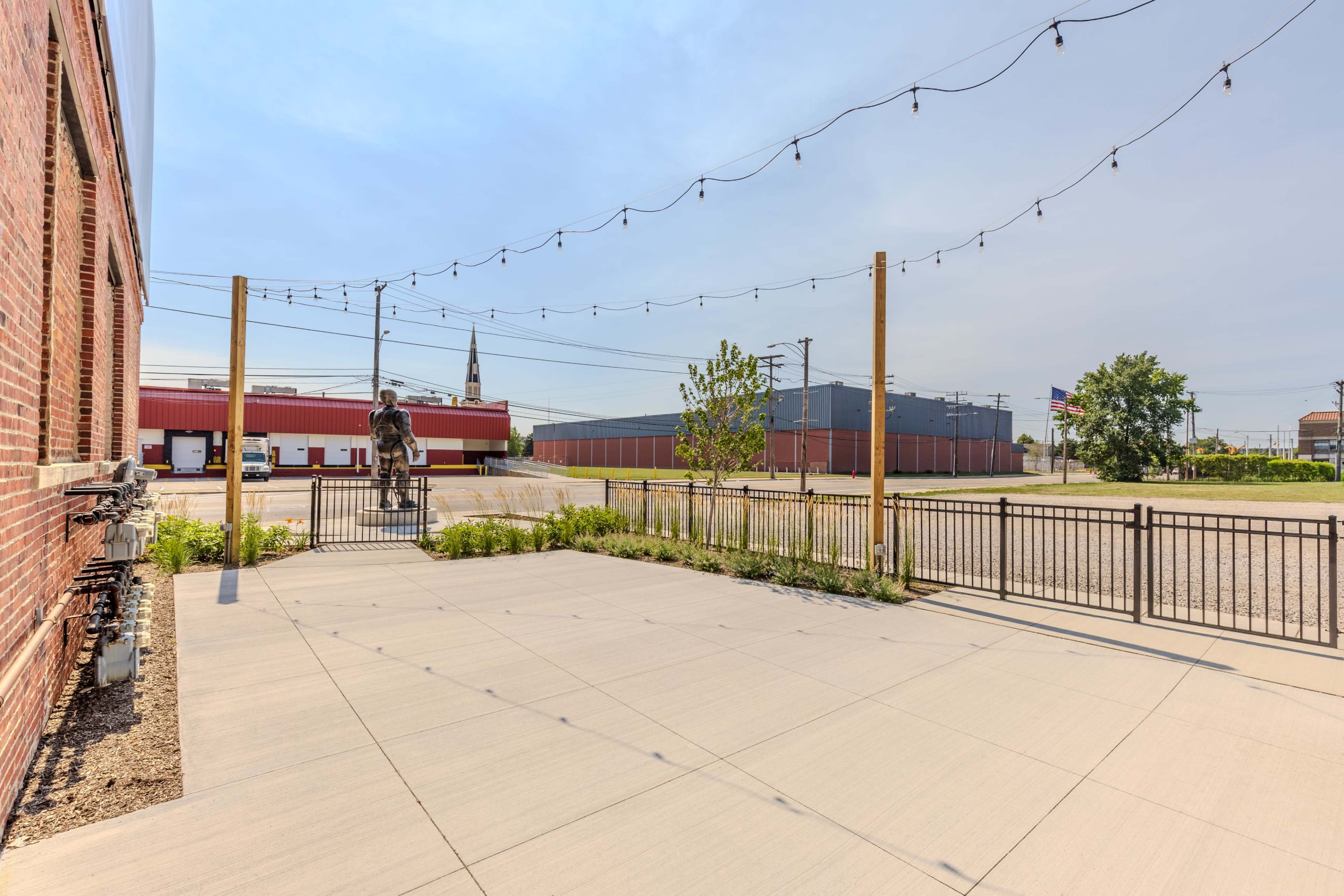 The image shows a fenced, paved outdoor area with string lights, a statue of a person on horseback, and a view of industrial buildings in the background.