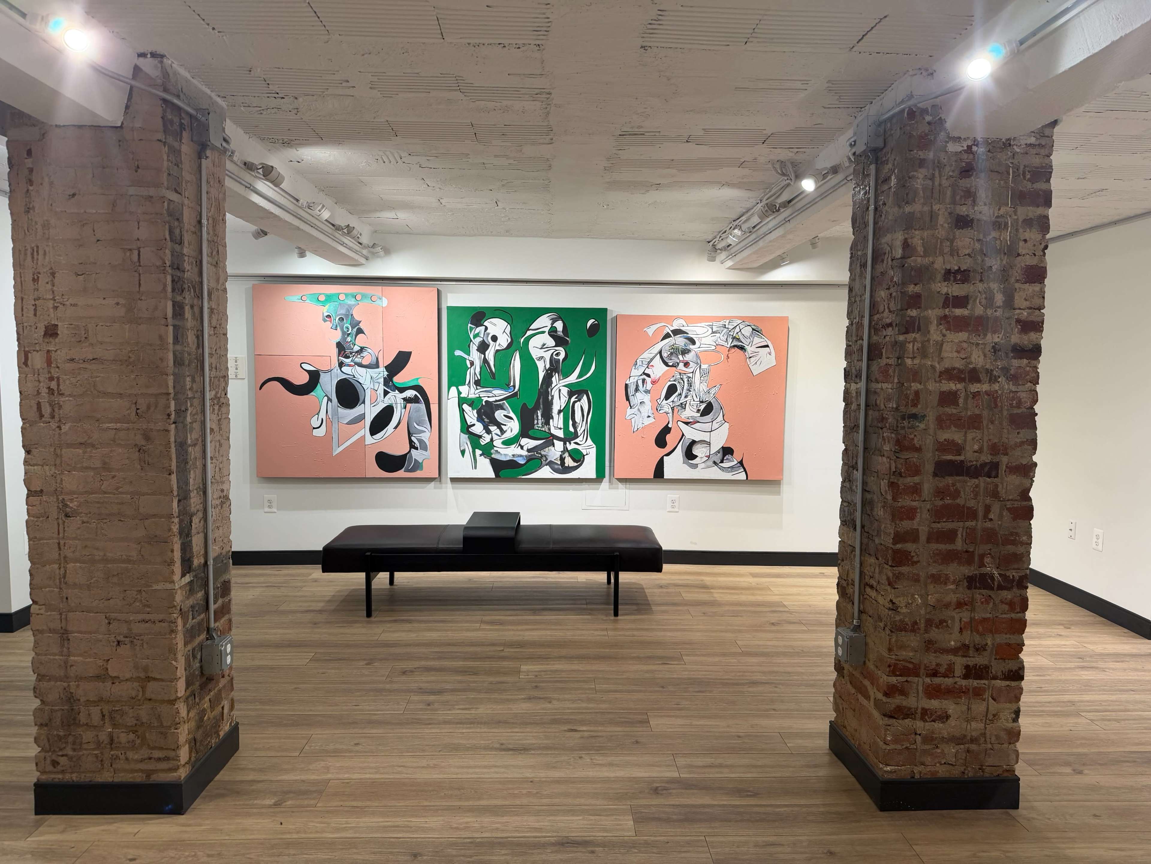 The image shows a contemporary art gallery featuring two large abstract paintings on the walls, with a brown bench in the center and exposed brick columns supporting the space.