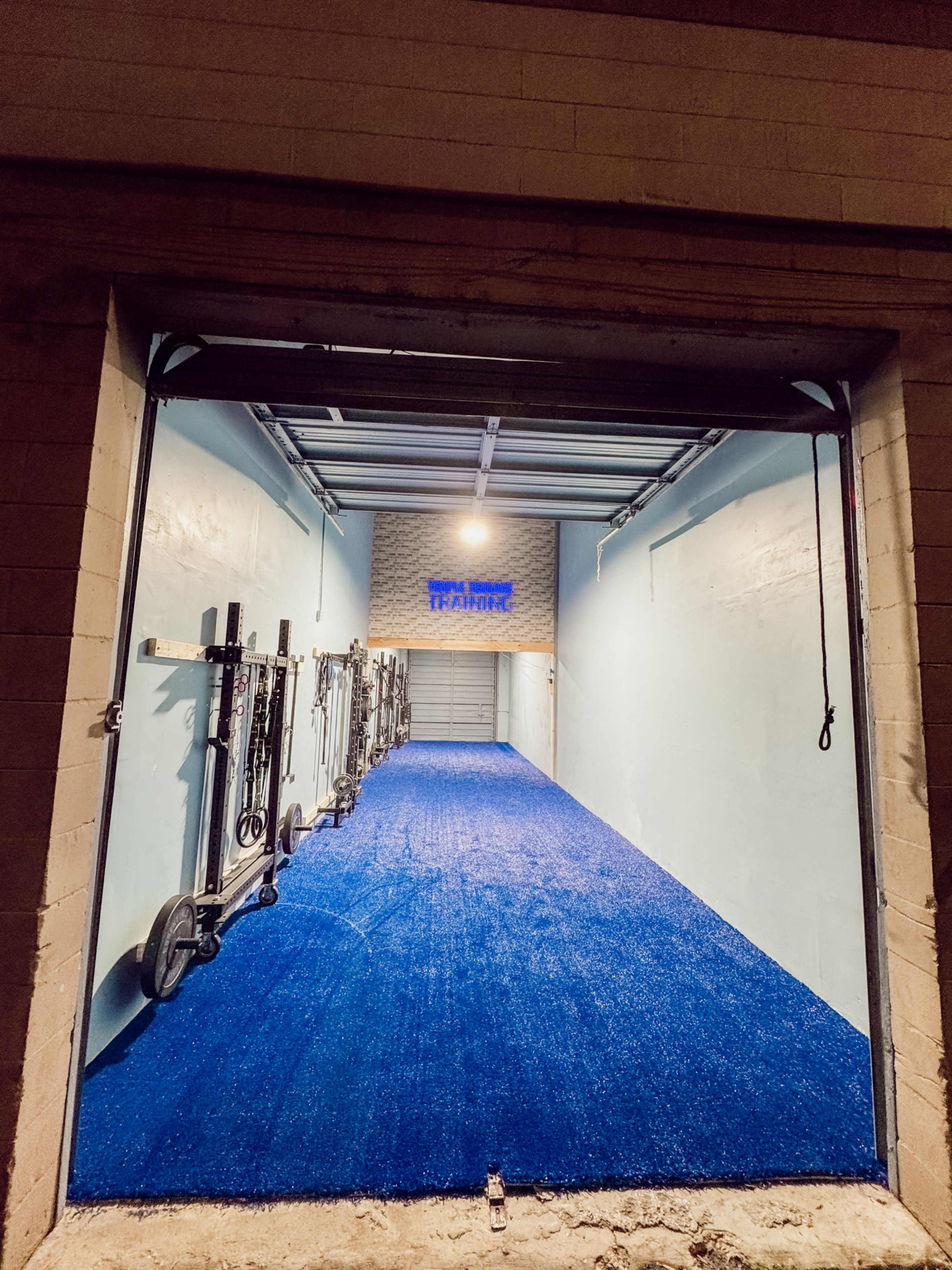 The image shows a training facility with a bright blue artificial turf floor and various gym equipment stored along the walls.