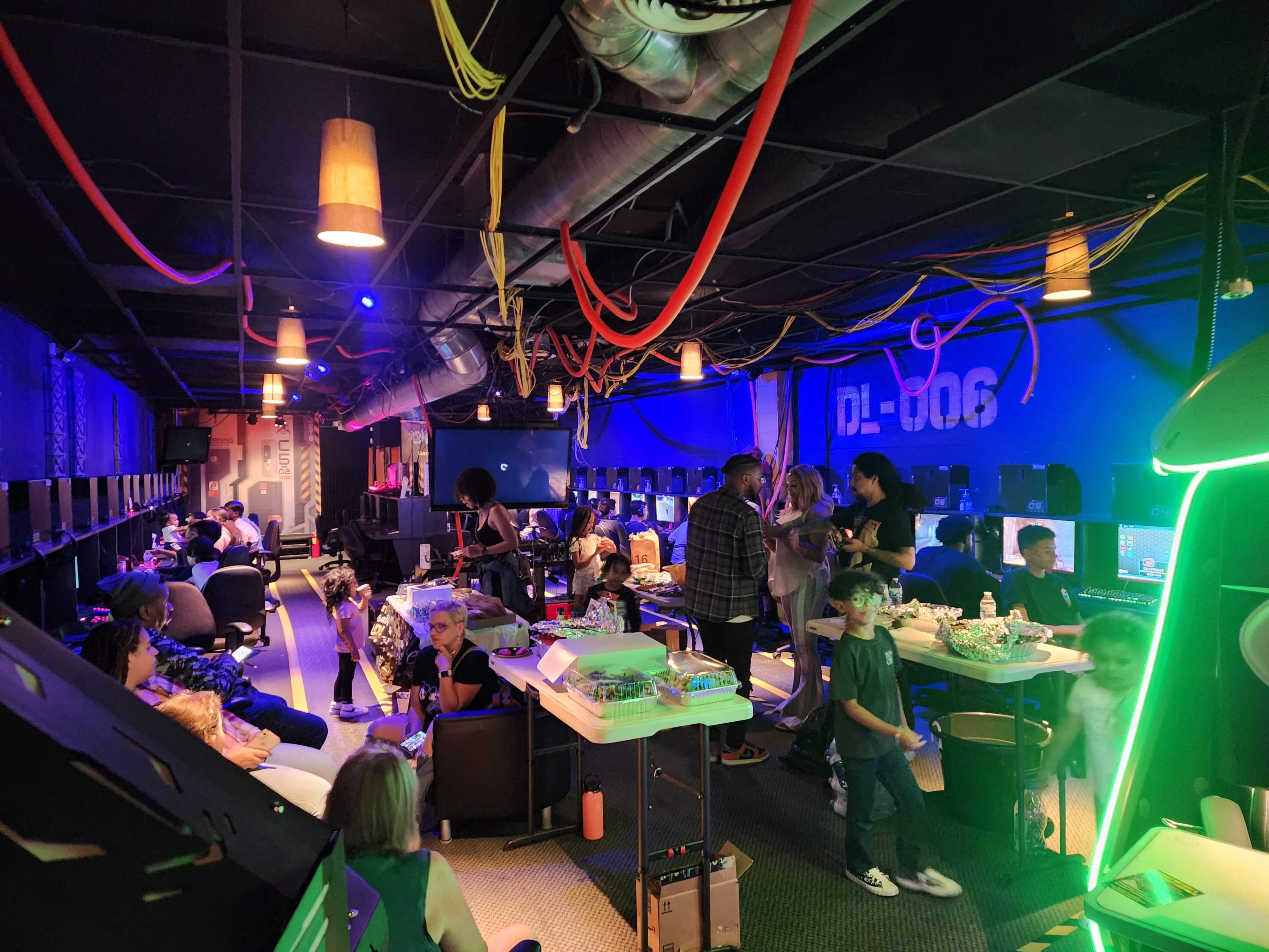 The image shows a crowded gaming lounge with people sitting and standing around tables, gaming stations lined up against the walls, and colorful lighting illuminating the space.