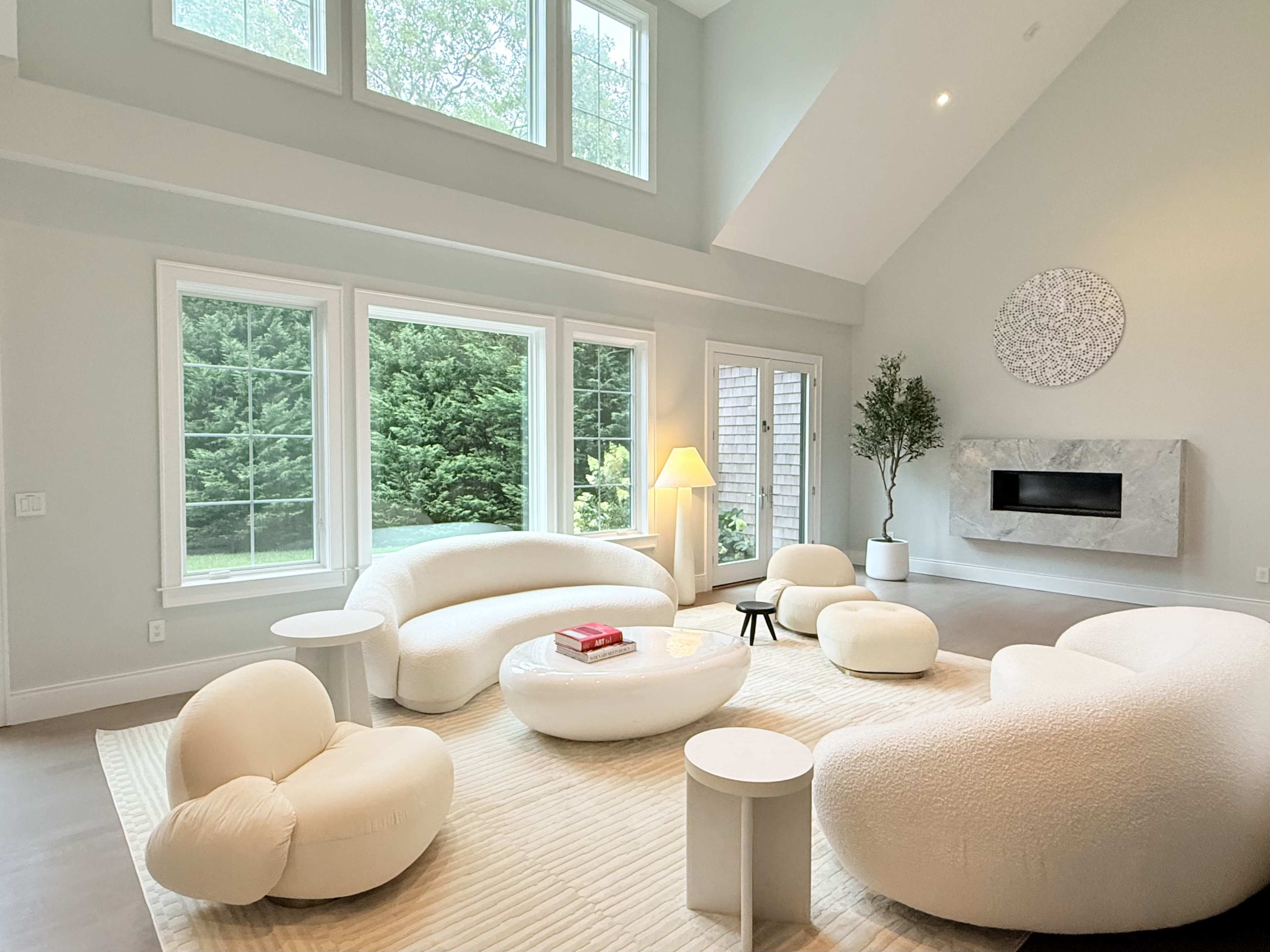 The image depicts a modern living room with curved, white furniture arranged around a low coffee table, large windows providing natural light, and a minimalist fireplace on the wall.
