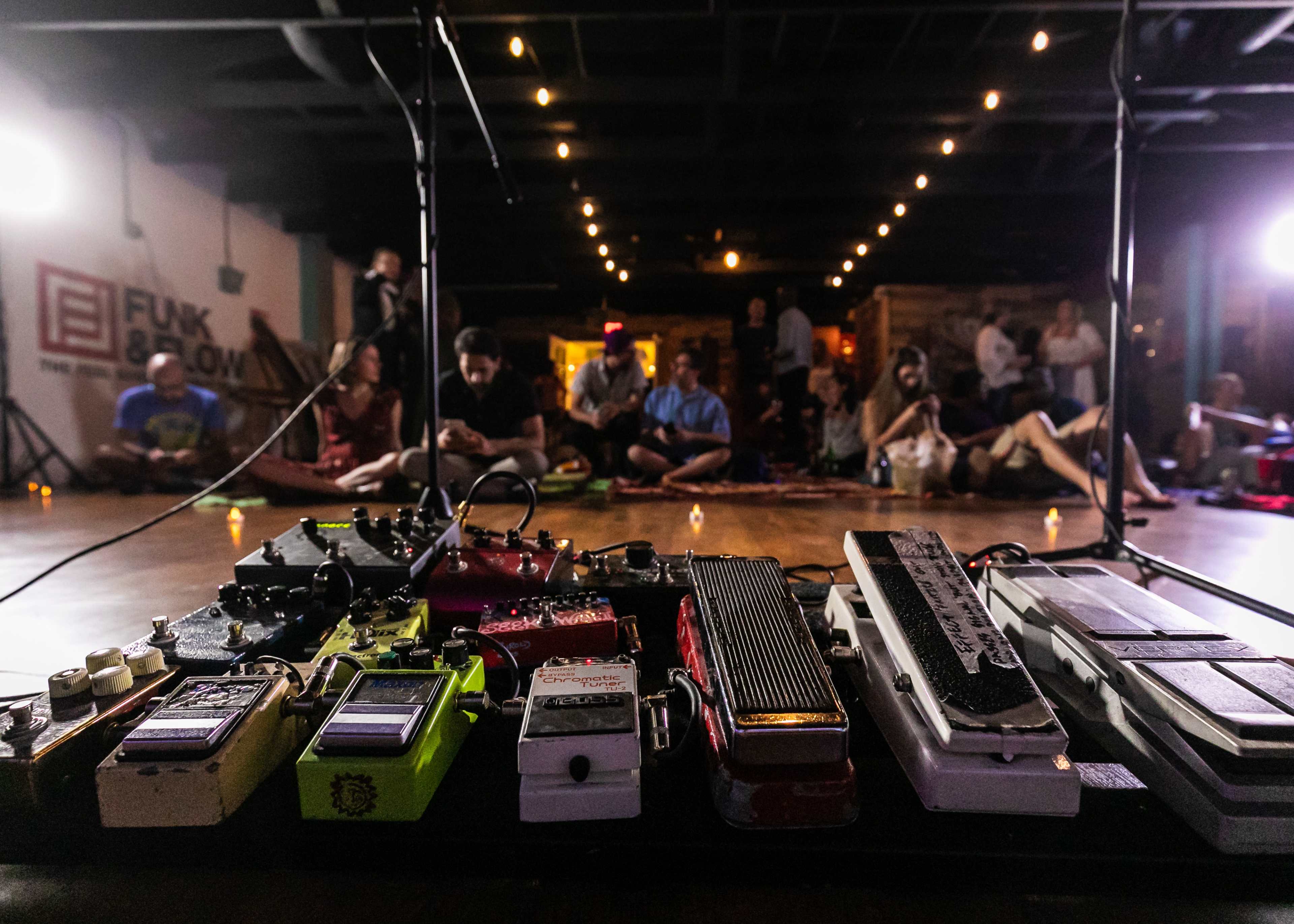 A collection of guitar effects pedals is prominently displayed in the foreground, with an audience seated in a dimly lit venue in the background.
