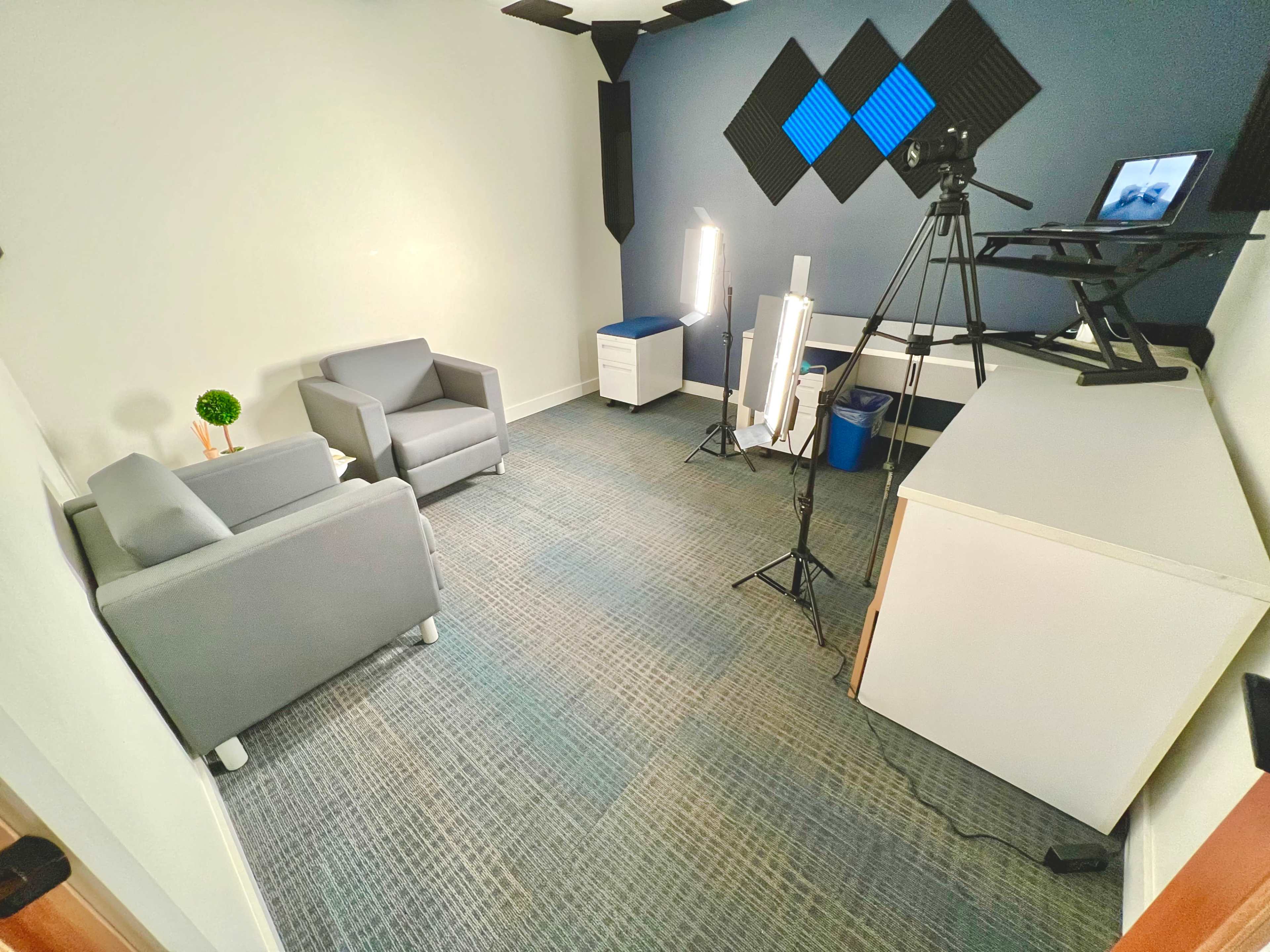 A small, empty room equipped with two gray chairs, a desk, and a camera setup on a tripod, with acoustic panels on the wall.