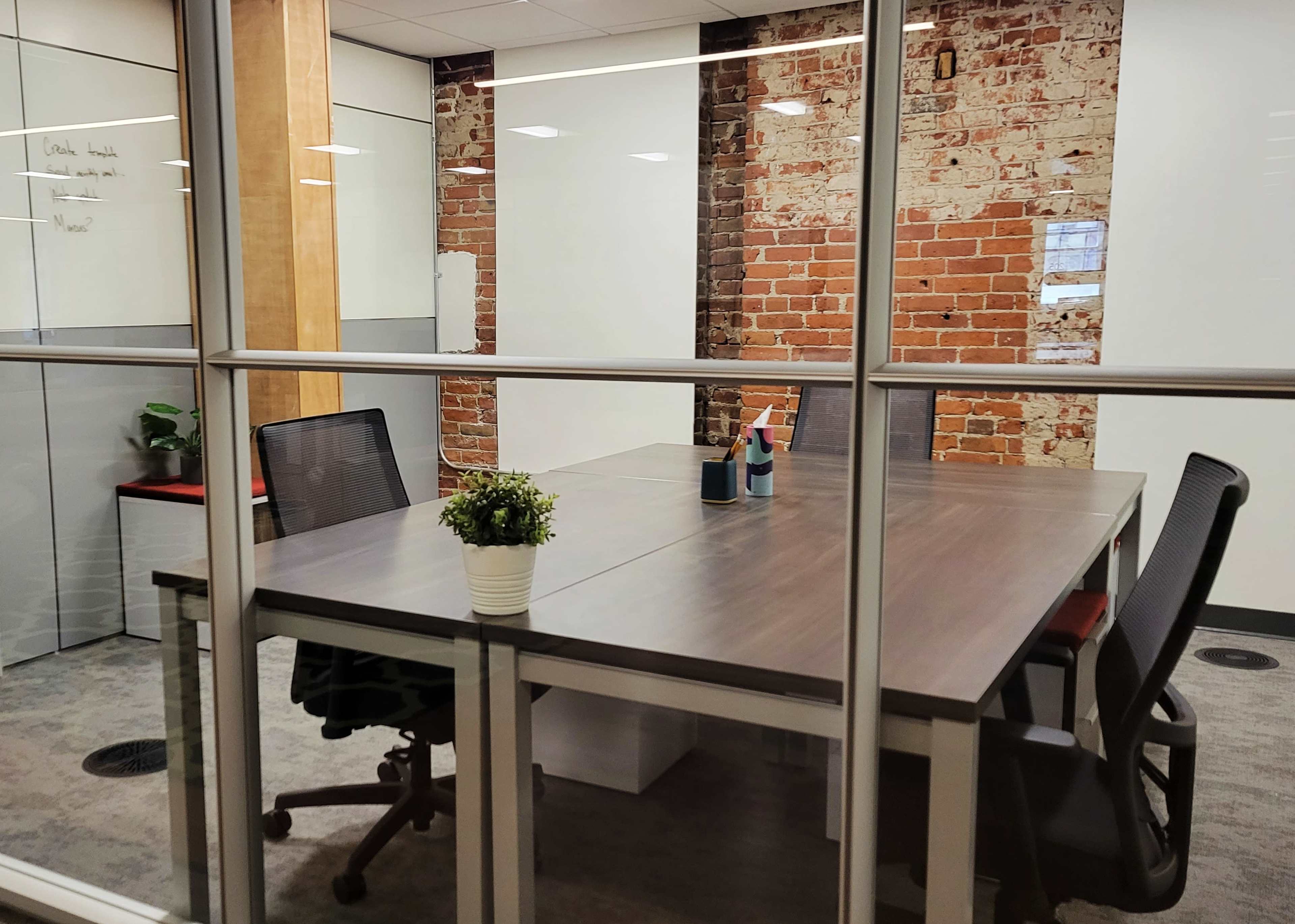 A conference room features a large table surrounded by chairs, with brick walls and plants visible through glass partitions.