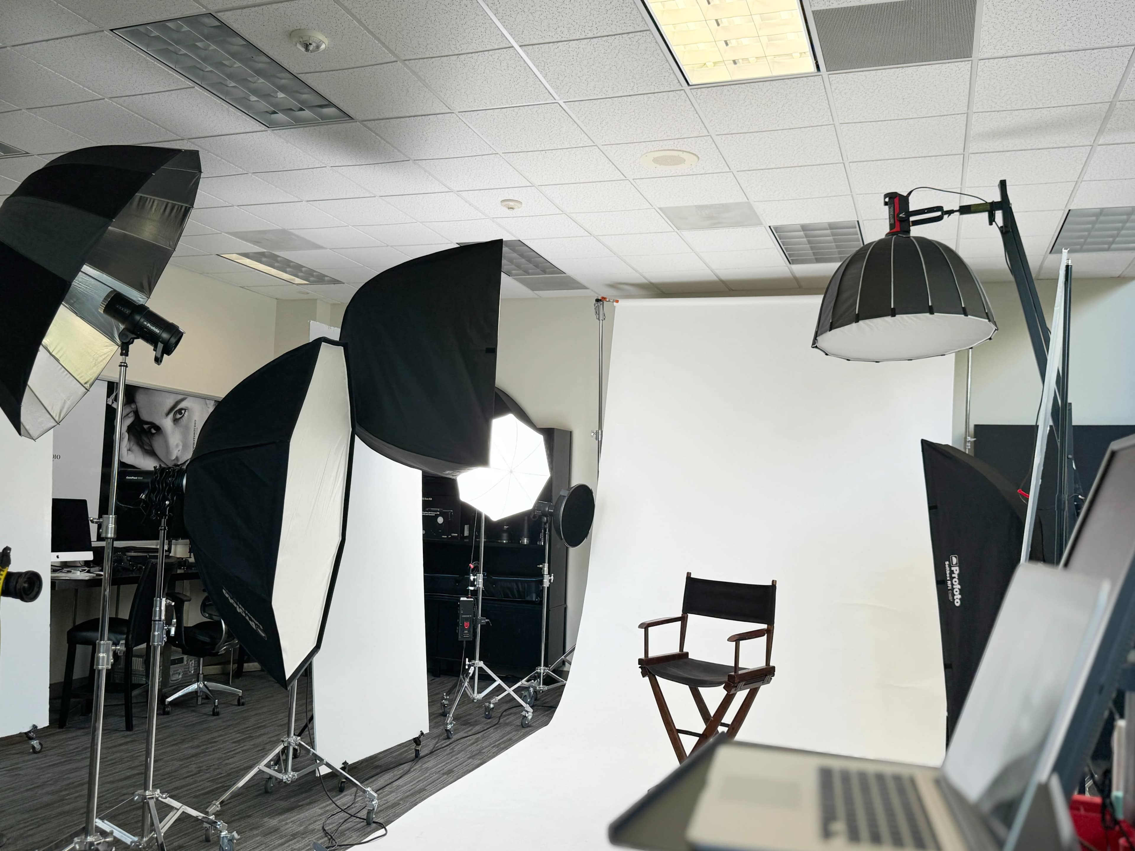 A photography studio is set up with various lights and reflectors aimed at a blank backdrop, featuring a director's chair in the center.