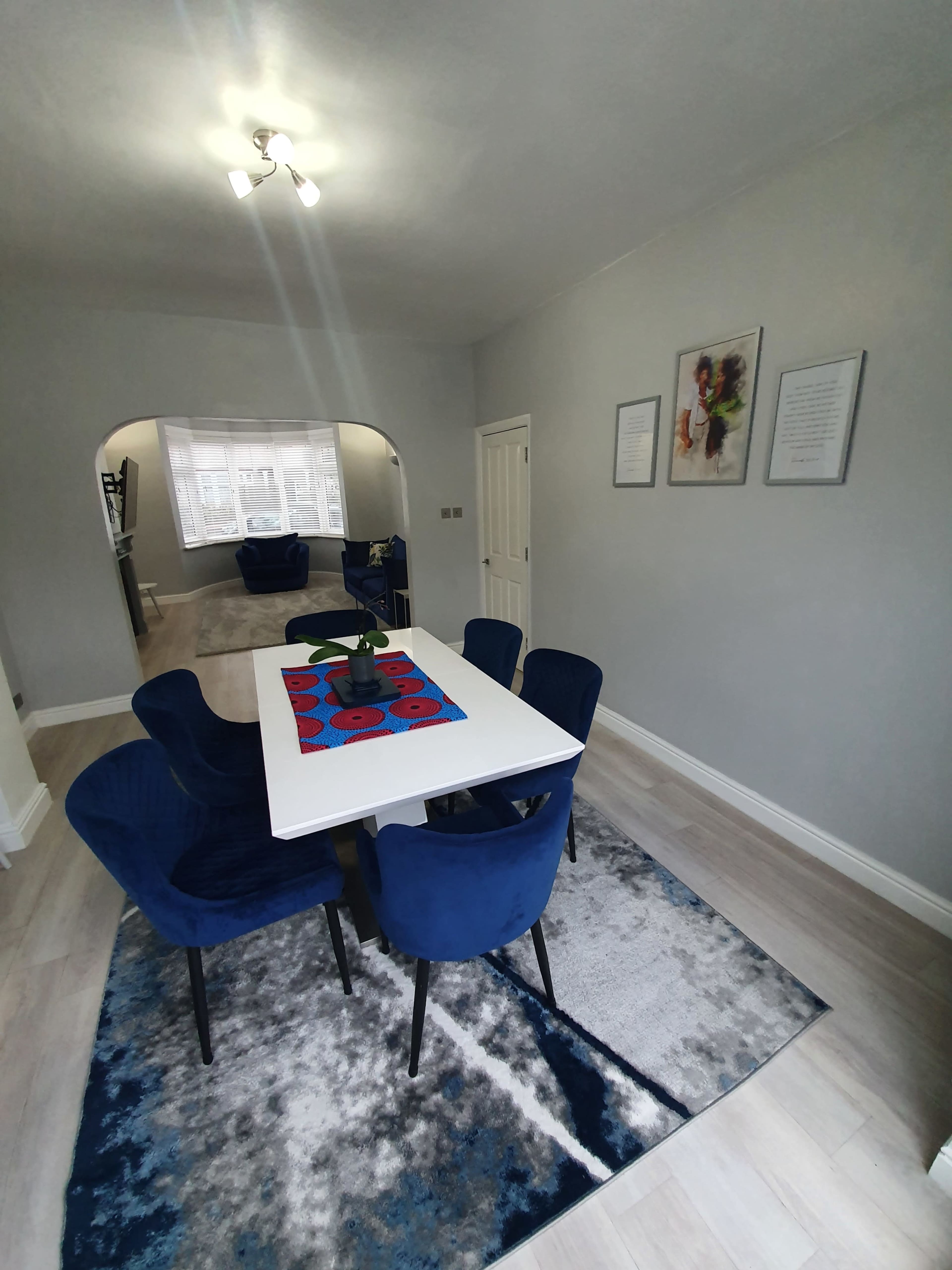 A dining area features a white table with a red and black centerpiece surrounded by blue upholstered chairs, with light gray walls and a light-colored floor.