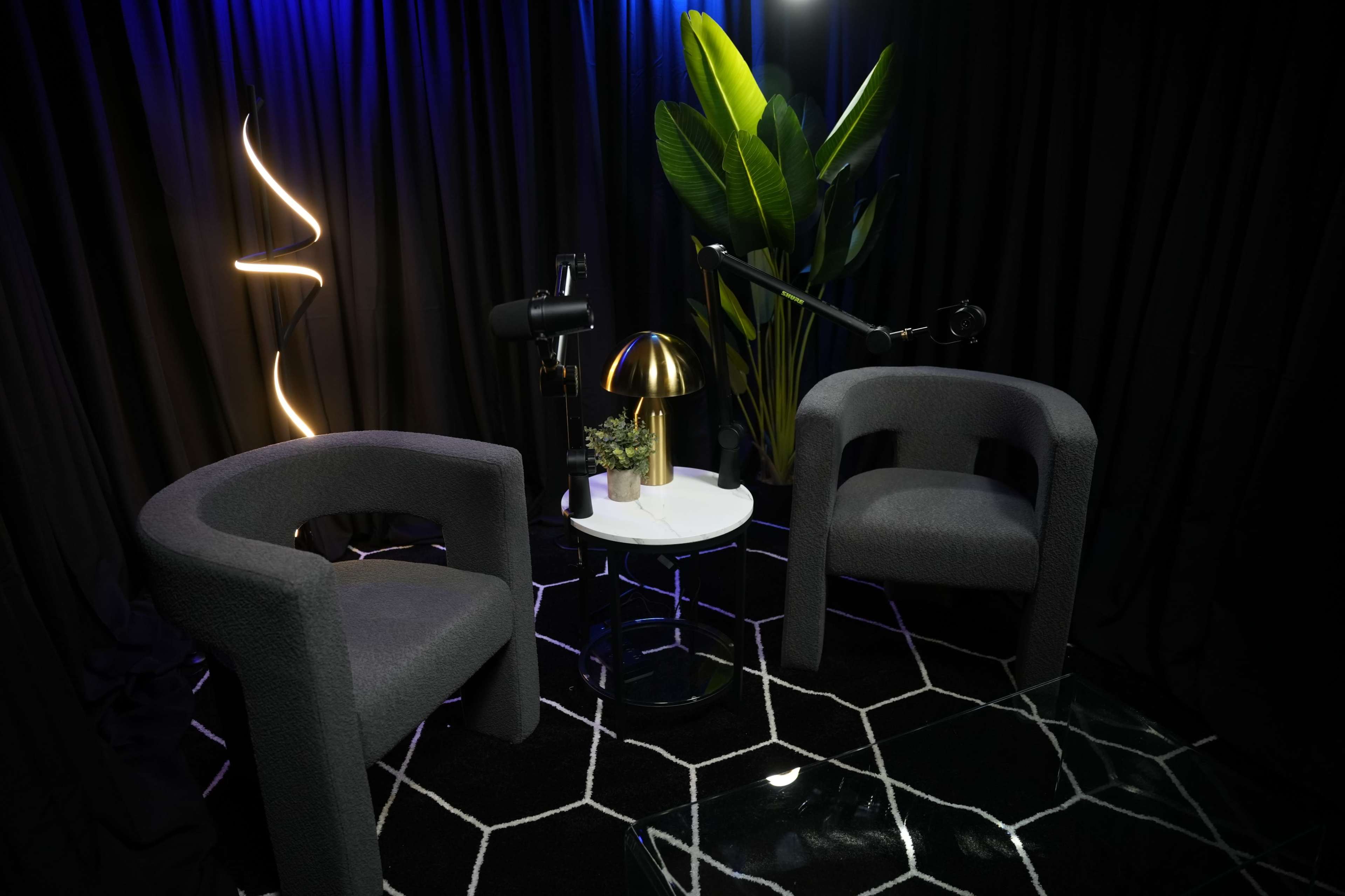 The image shows a small, modern conversation area with two gray chairs, a round table, a lamp, and decorative plants, set against a black curtain backdrop.