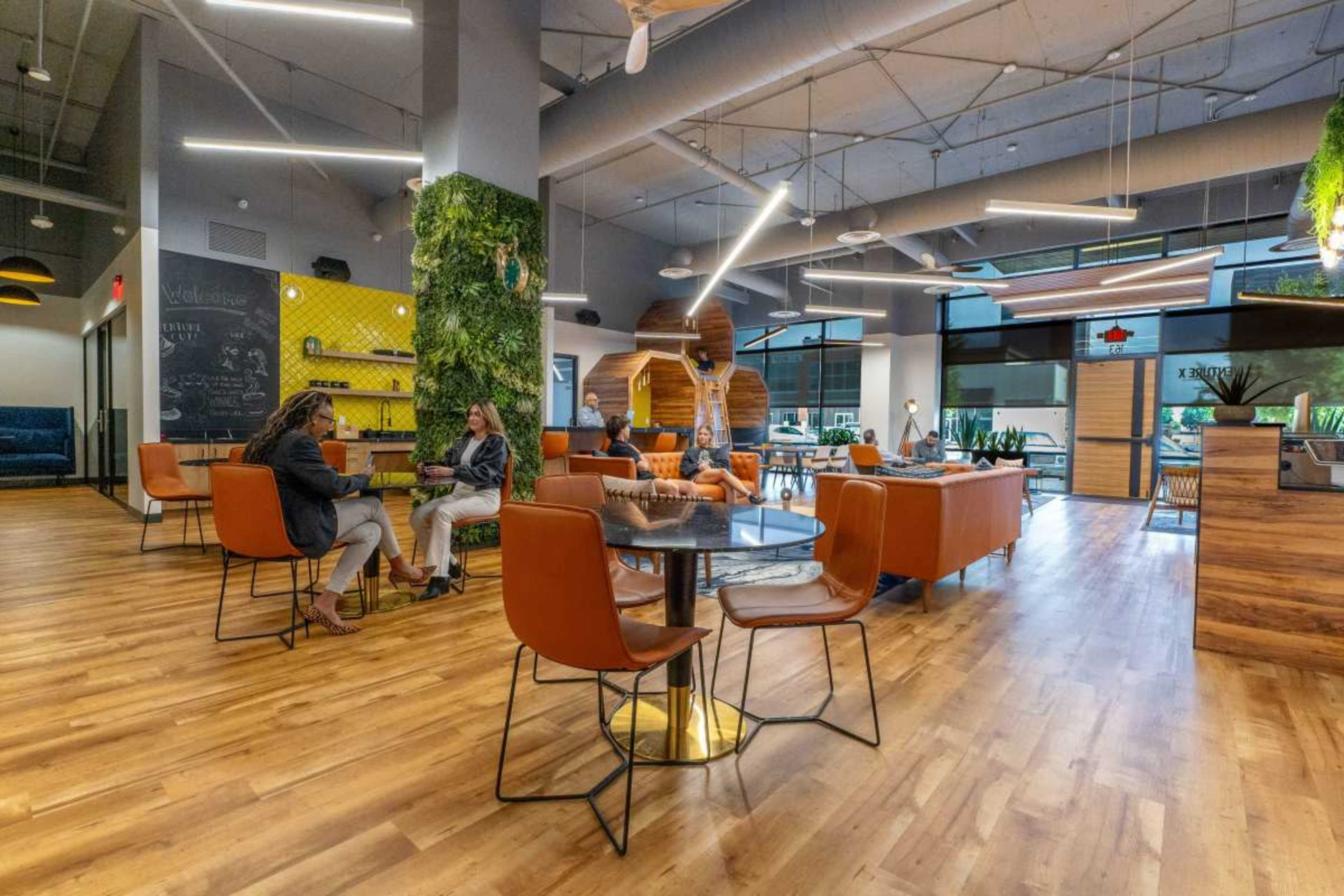 A modern lounge area features a mix of seating arrangements, greenery on the walls, and large windows allowing natural light.