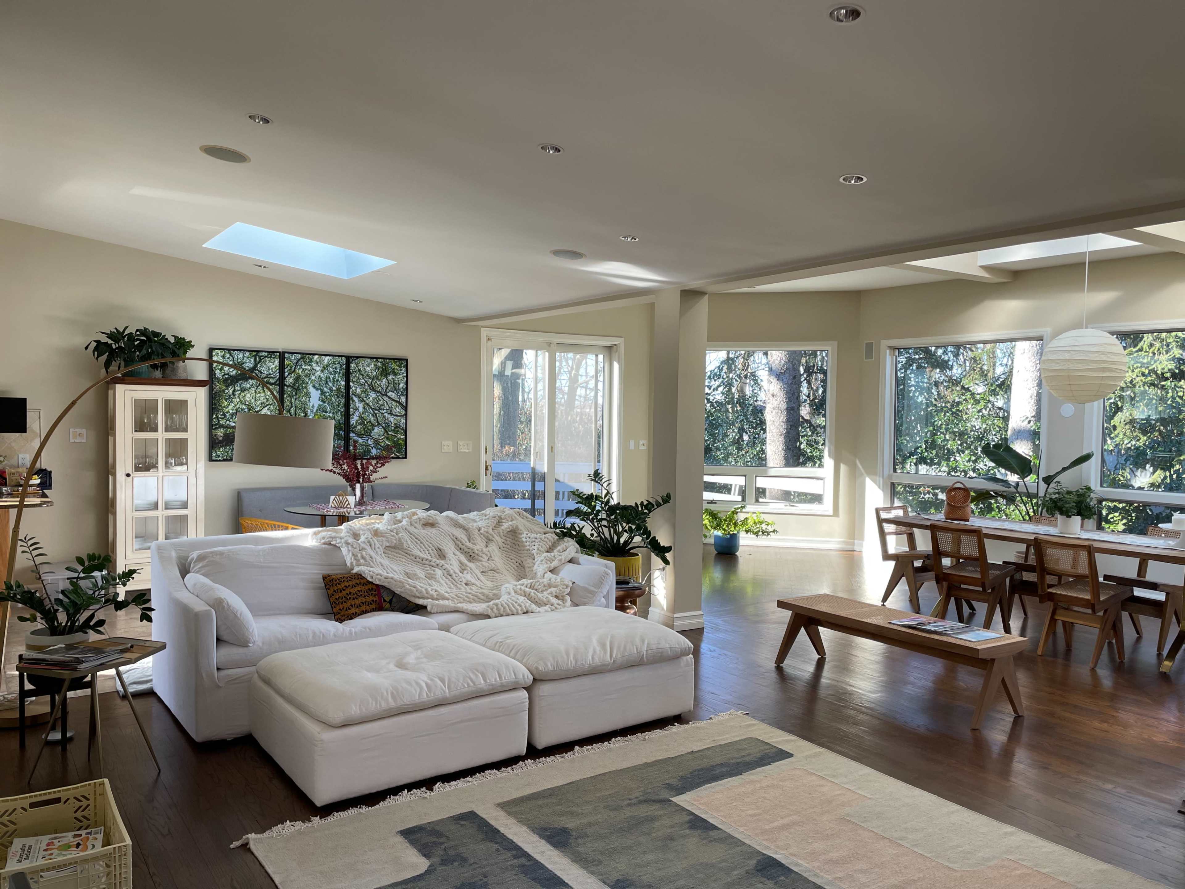 A bright, open living space features a white sectional sofa, wooden dining table, and large windows overlooking greenery.