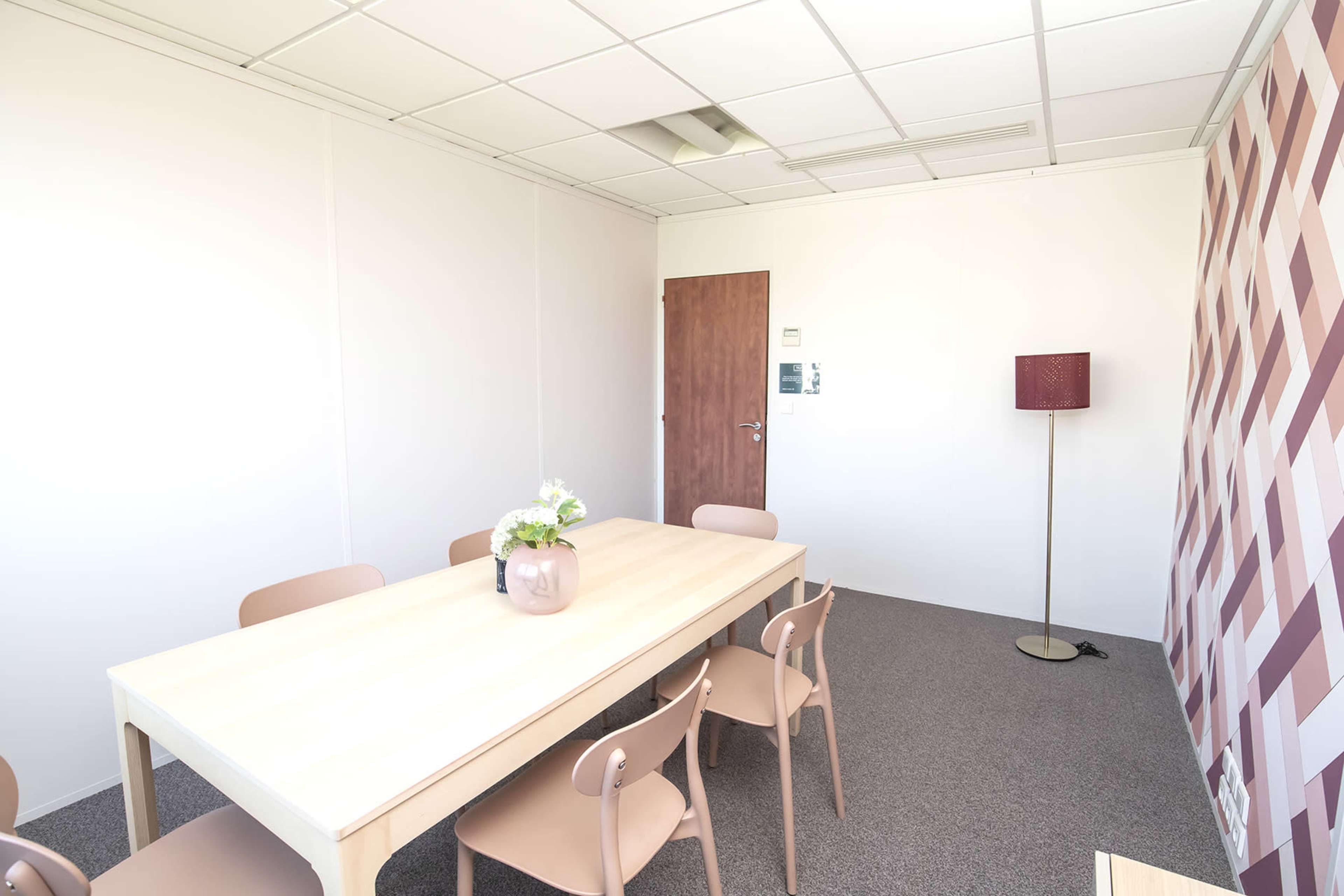 A modern meeting room with a long wooden table surrounded by beige chairs, a floor lamp, and a patterned accent wall.