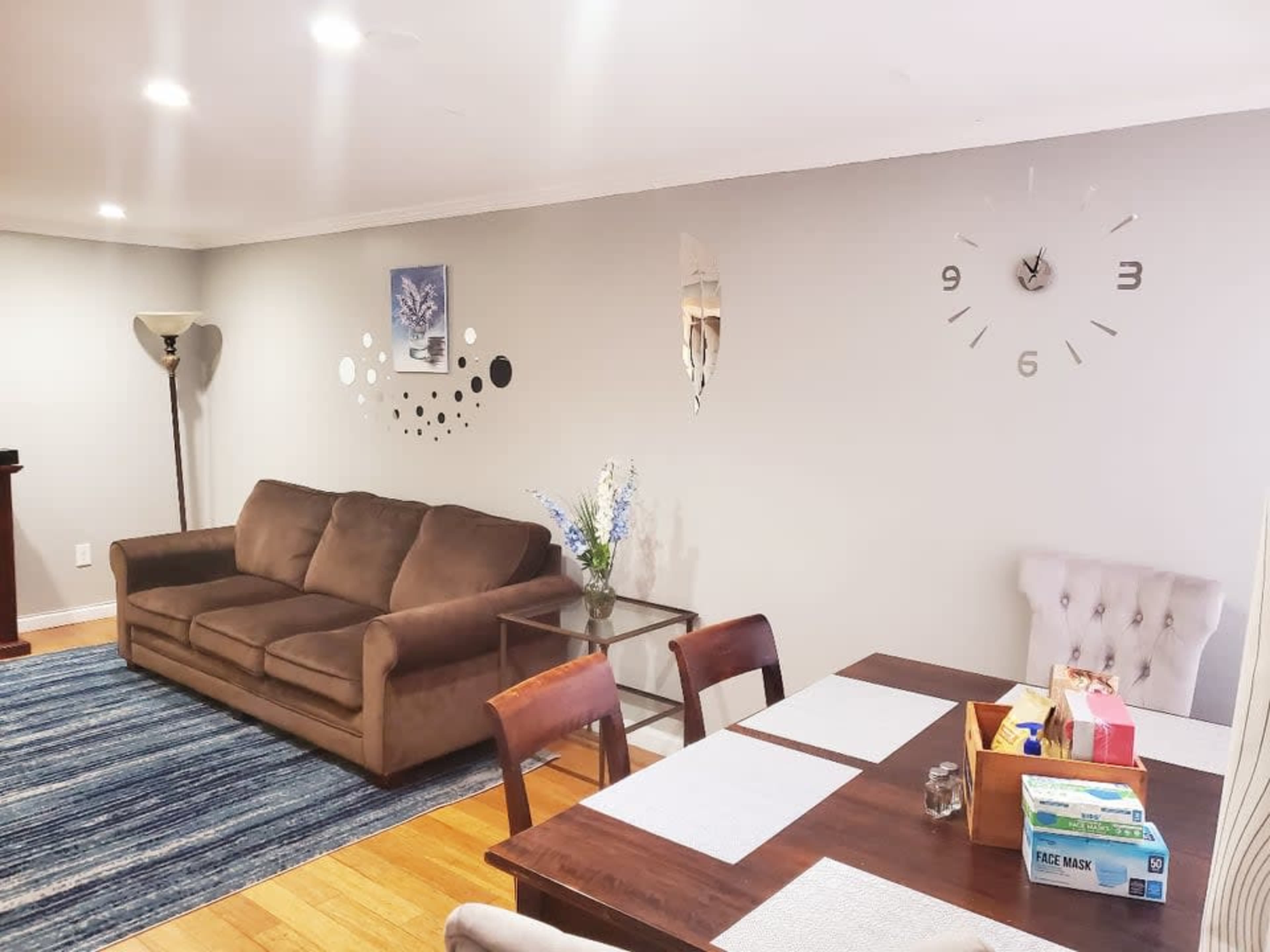 A living room with a brown sofa, a dining area with a wooden table and chairs, and a clock on the wall.