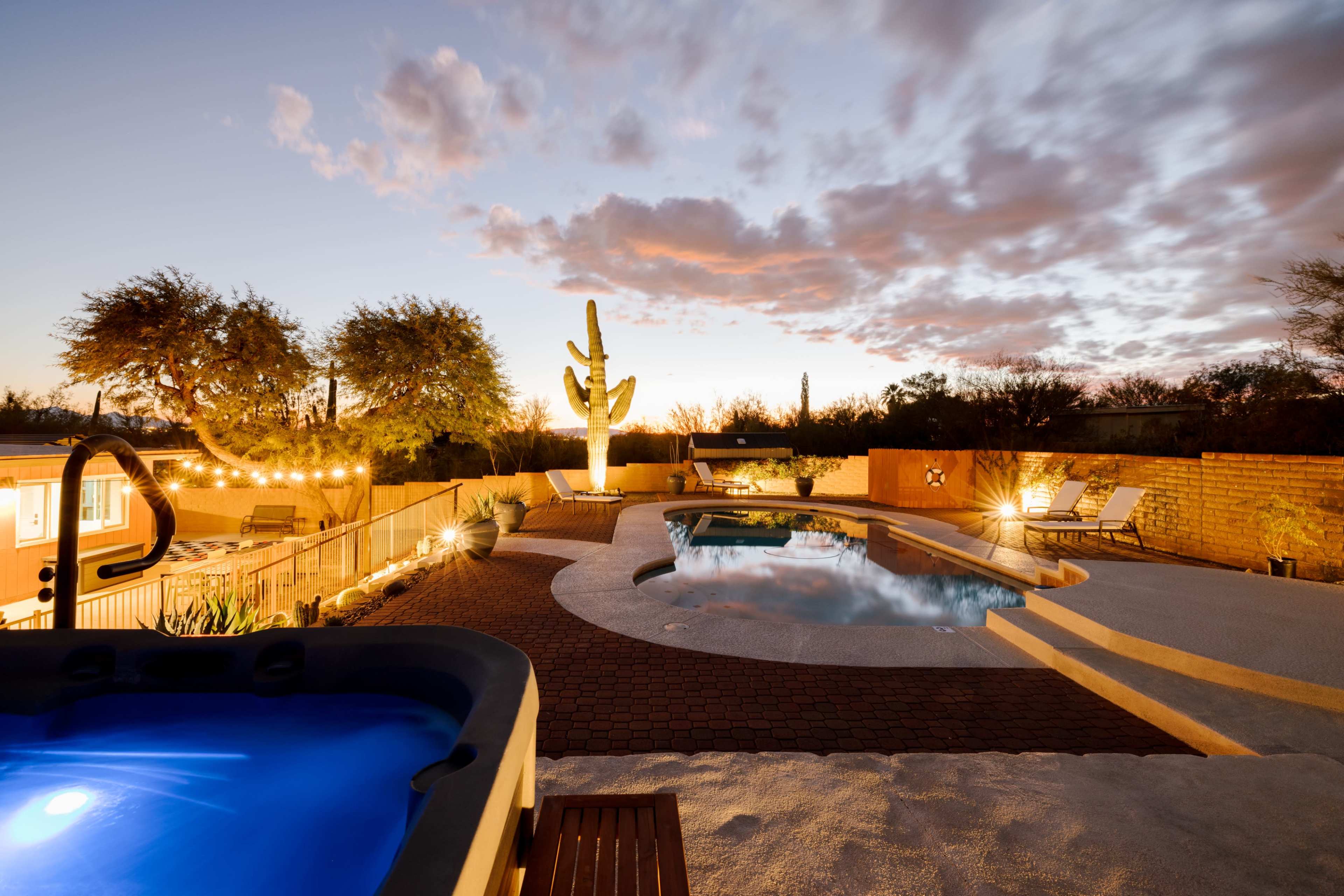 A desert backyard at dusk with a hot tub, swimming pool, and surrounding landscaping featuring a large cactus.