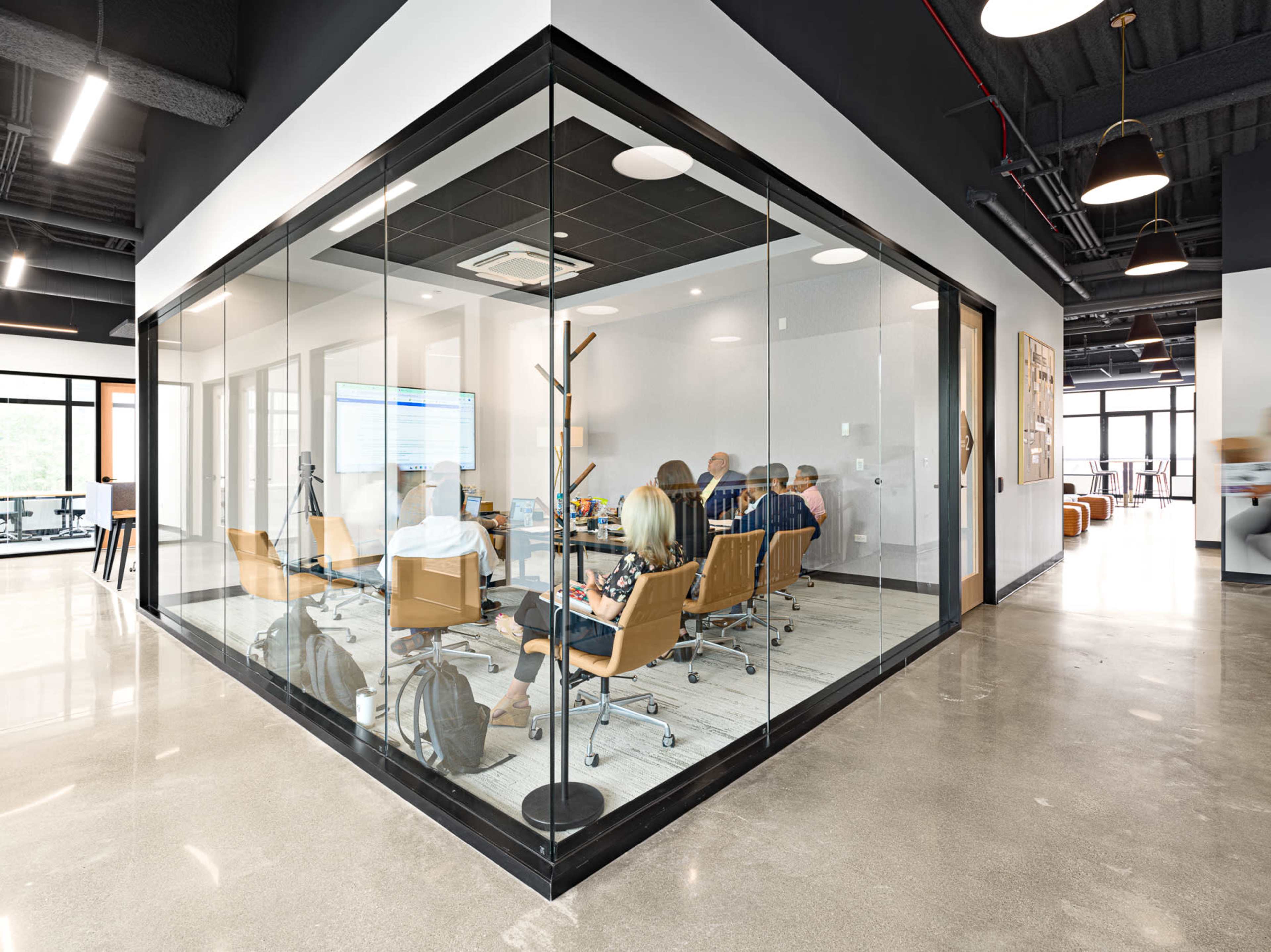 A group of people is seated around a conference table in a glass-walled meeting room.