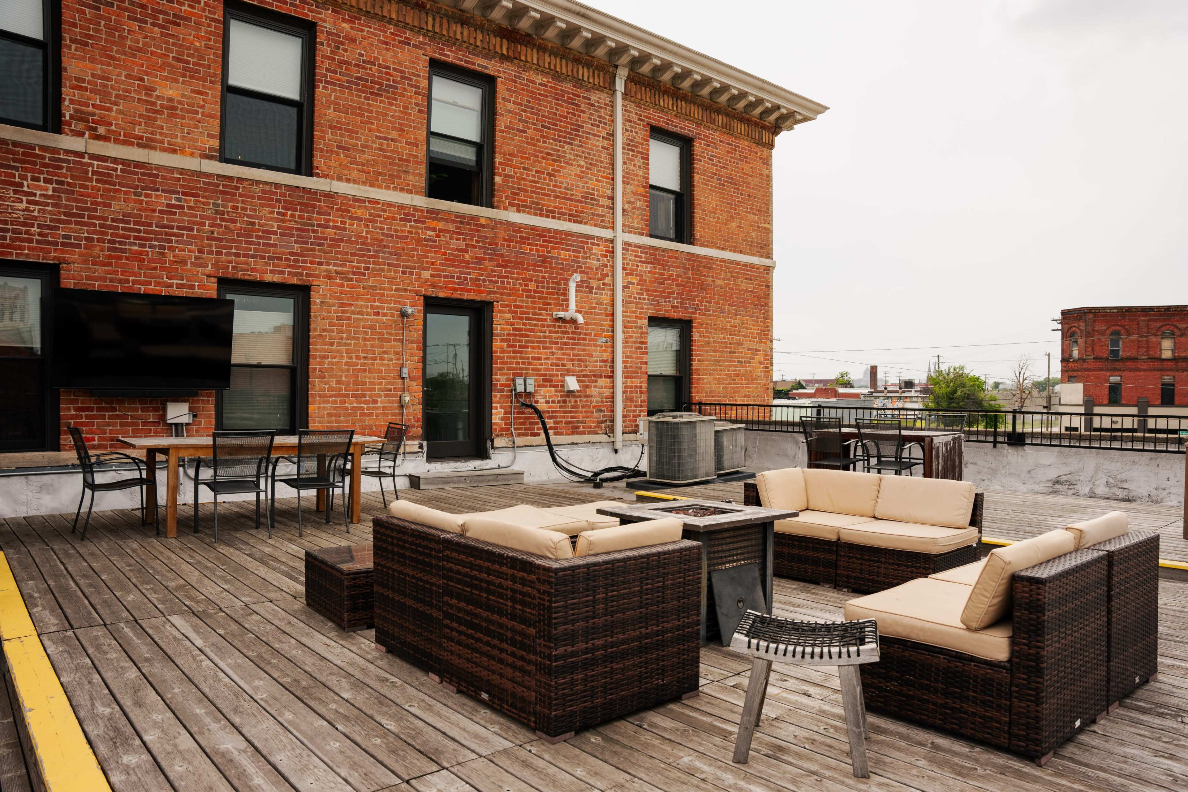 The image shows a rooftop patio with a seating area made of wicker furniture, a dining table, and a television mounted on a brick building.