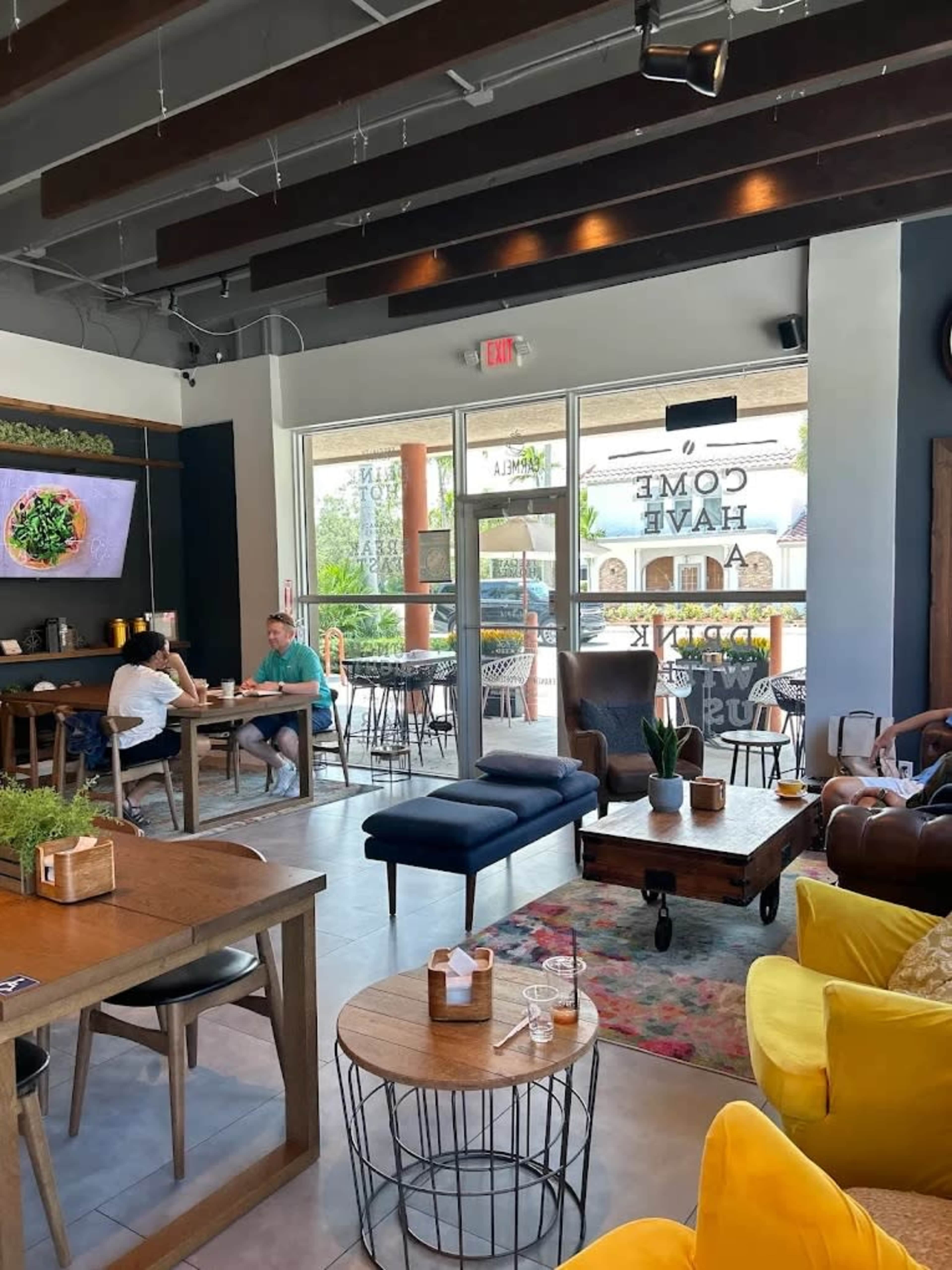 The image shows a modern café interior with various seating options, including a wooden table, a blue sofa, and yellow armchairs, along with a large window displaying outdoor views.