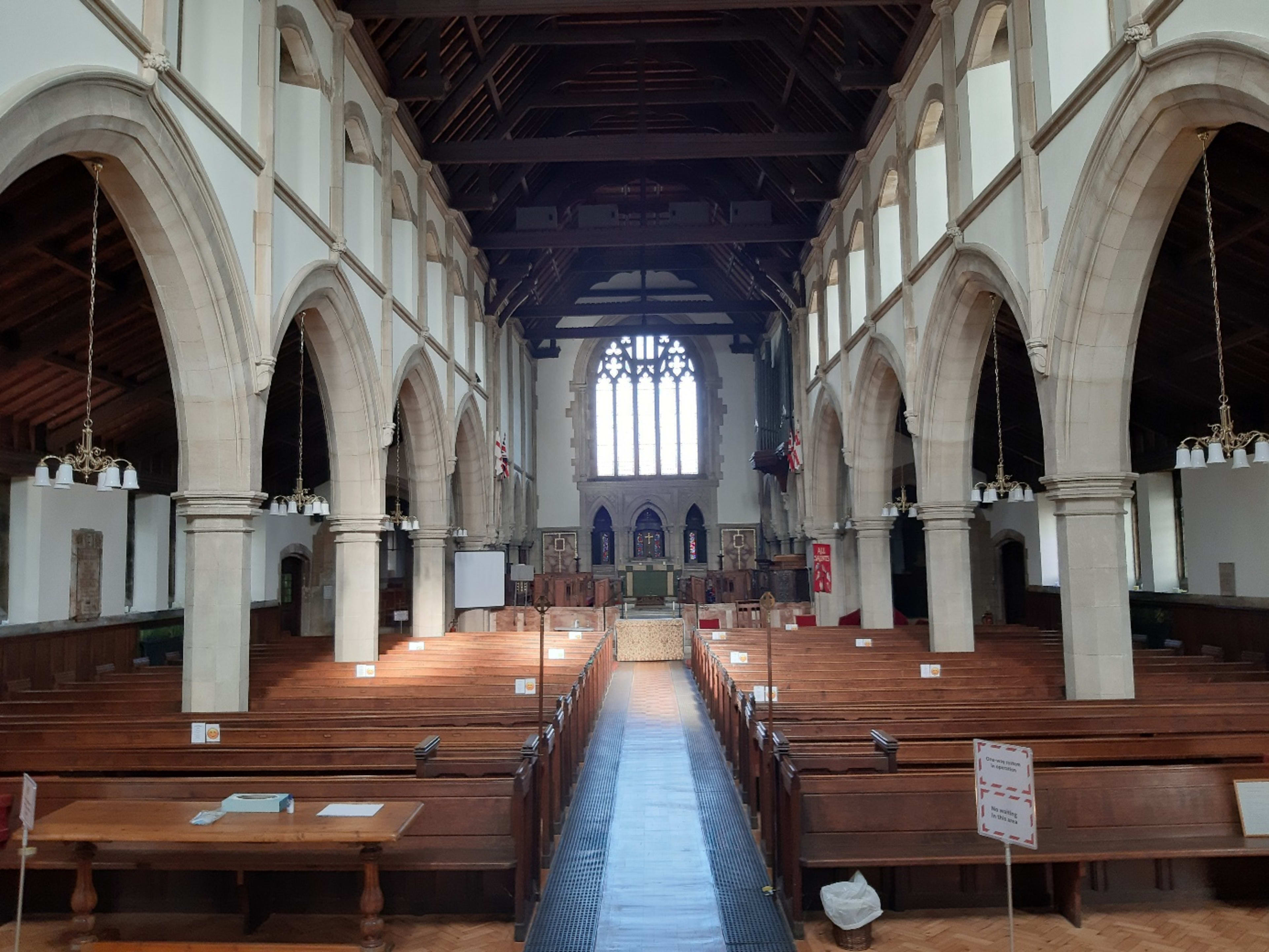 The interior of a large church featuring wooden pews arranged in rows, arched stone columns, and a high, beamed ceiling with a large stained glass window at one end.