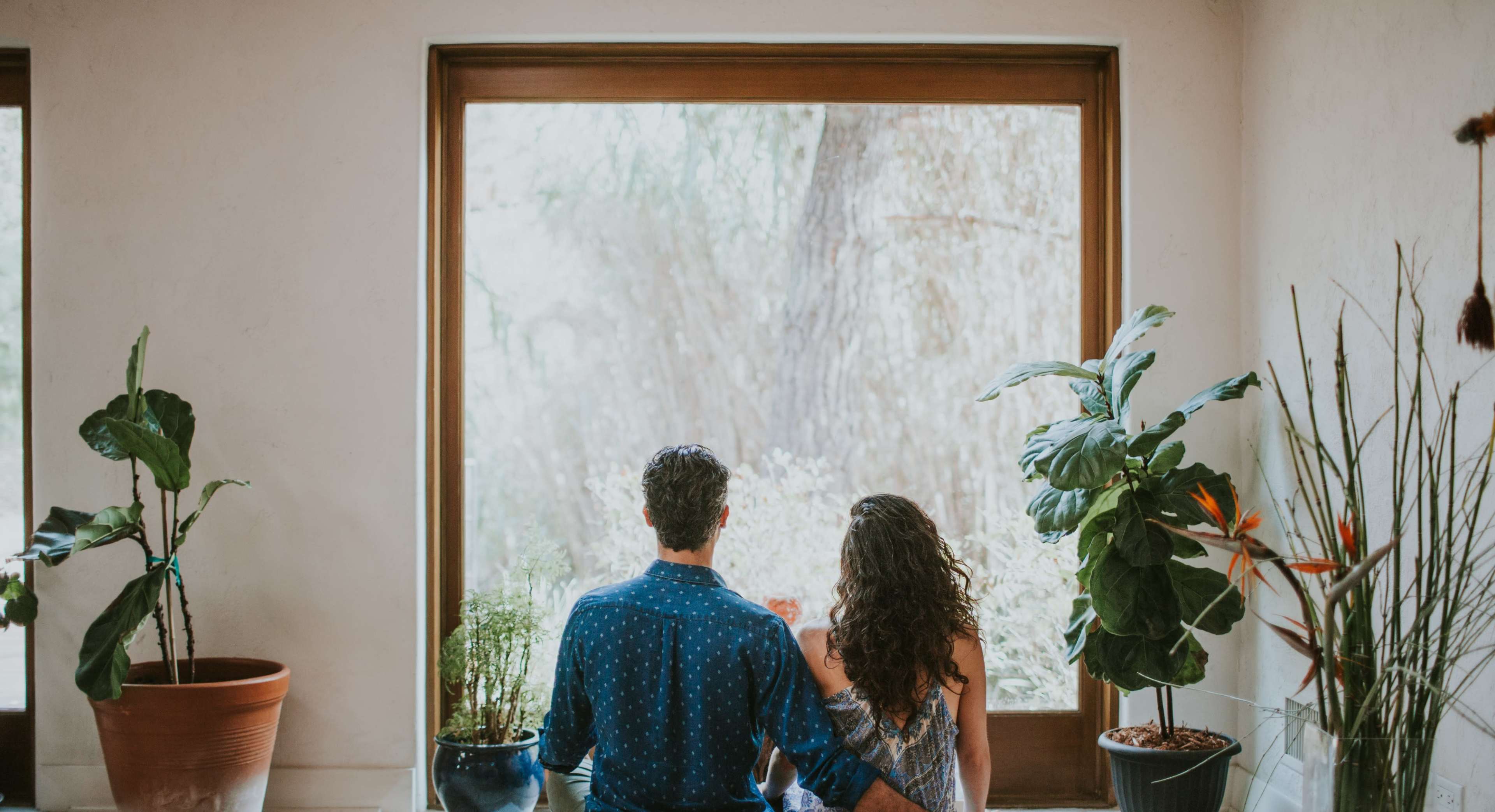 A man and a woman sit side by side, facing a large window with plants in pots, looking out at a natural scene.
