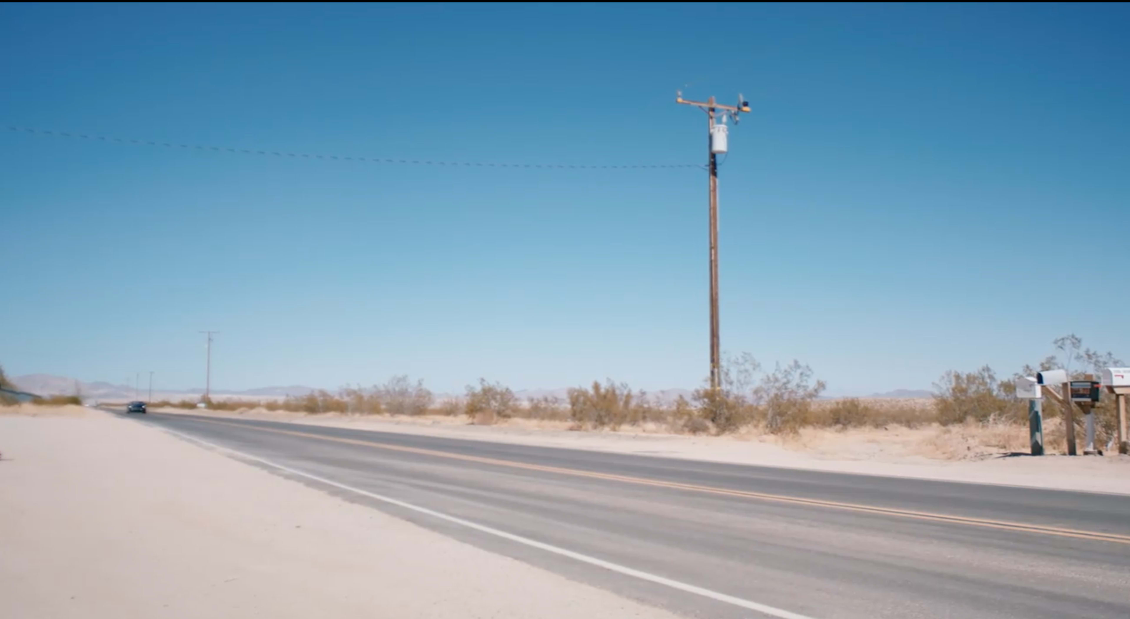 A long, empty road stretches across a desert landscape under a clear blue sky, with a power pole and sparse vegetation along the sides.