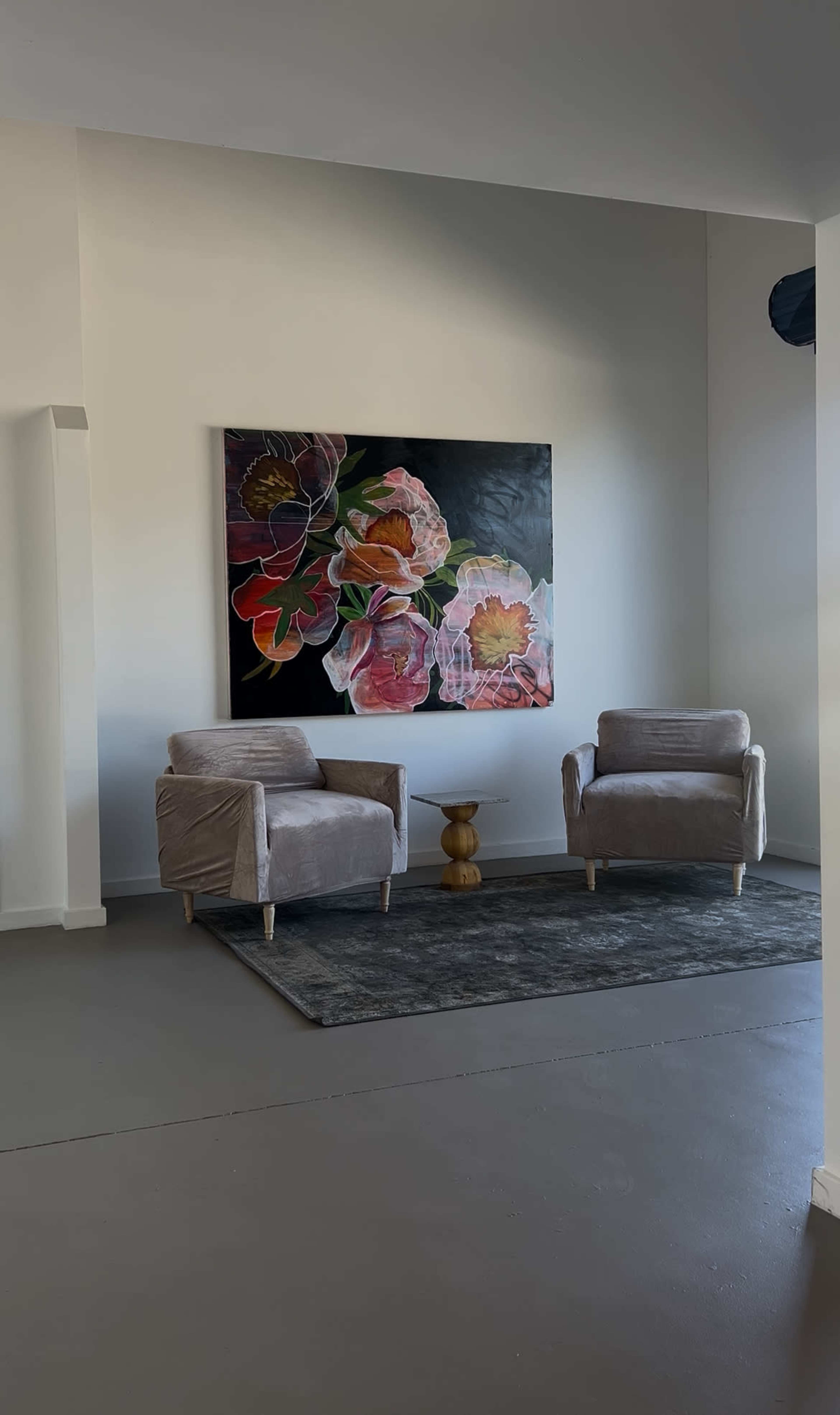 The scene features two gray upholstered chairs positioned on a rug in front of a large floral painting mounted on a white wall.
