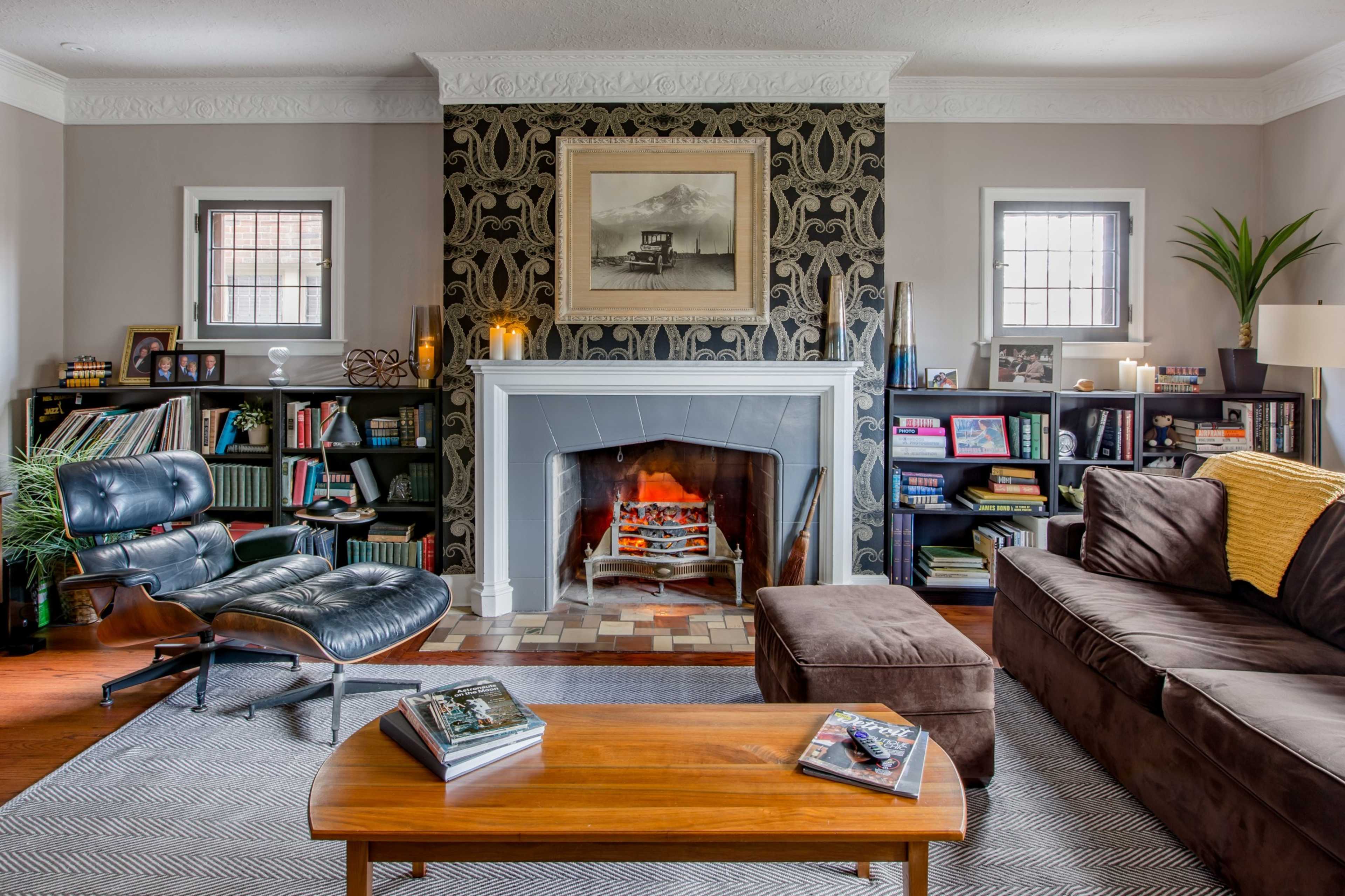 A cozy living room with a fireplace, bookshelves filled with books and decorative items, a large brown sofa, an armchair, and a wooden coffee table.