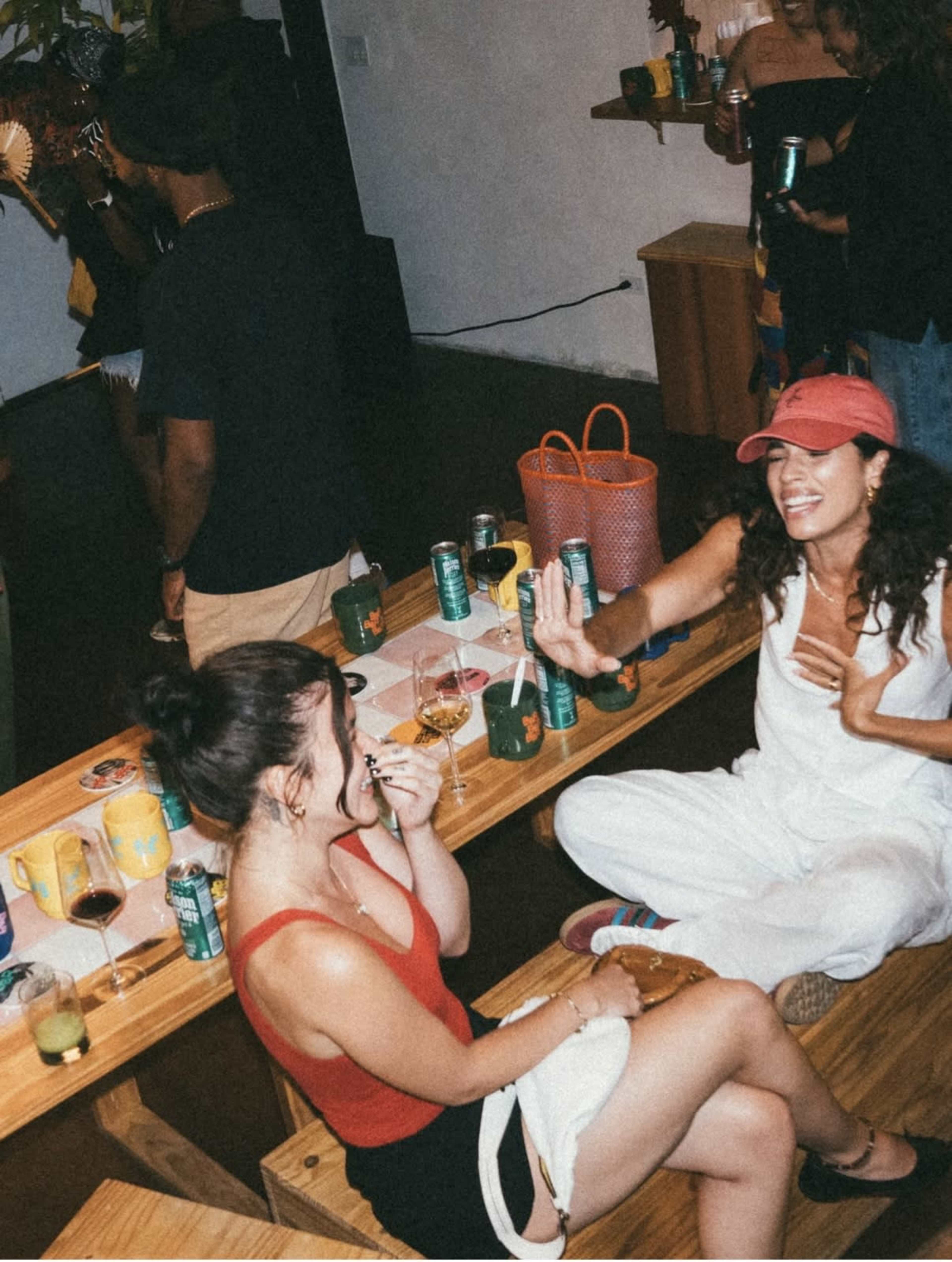 Two women are laughing while sitting at a wooden table covered with drinks and snacks, as others socialize in the background.