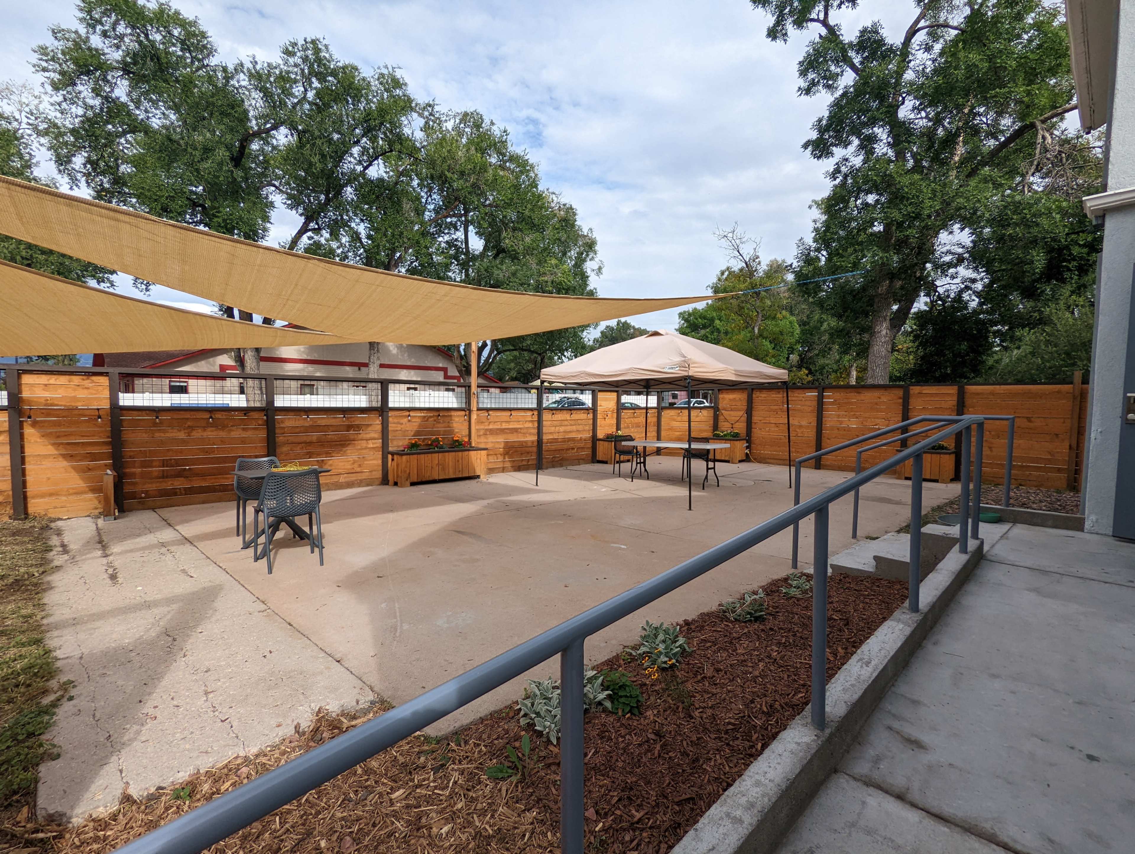A patio area with a concrete floor, surrounded by wooden fencing, featuring two seated areas under shade sails and a table with an umbrella.
