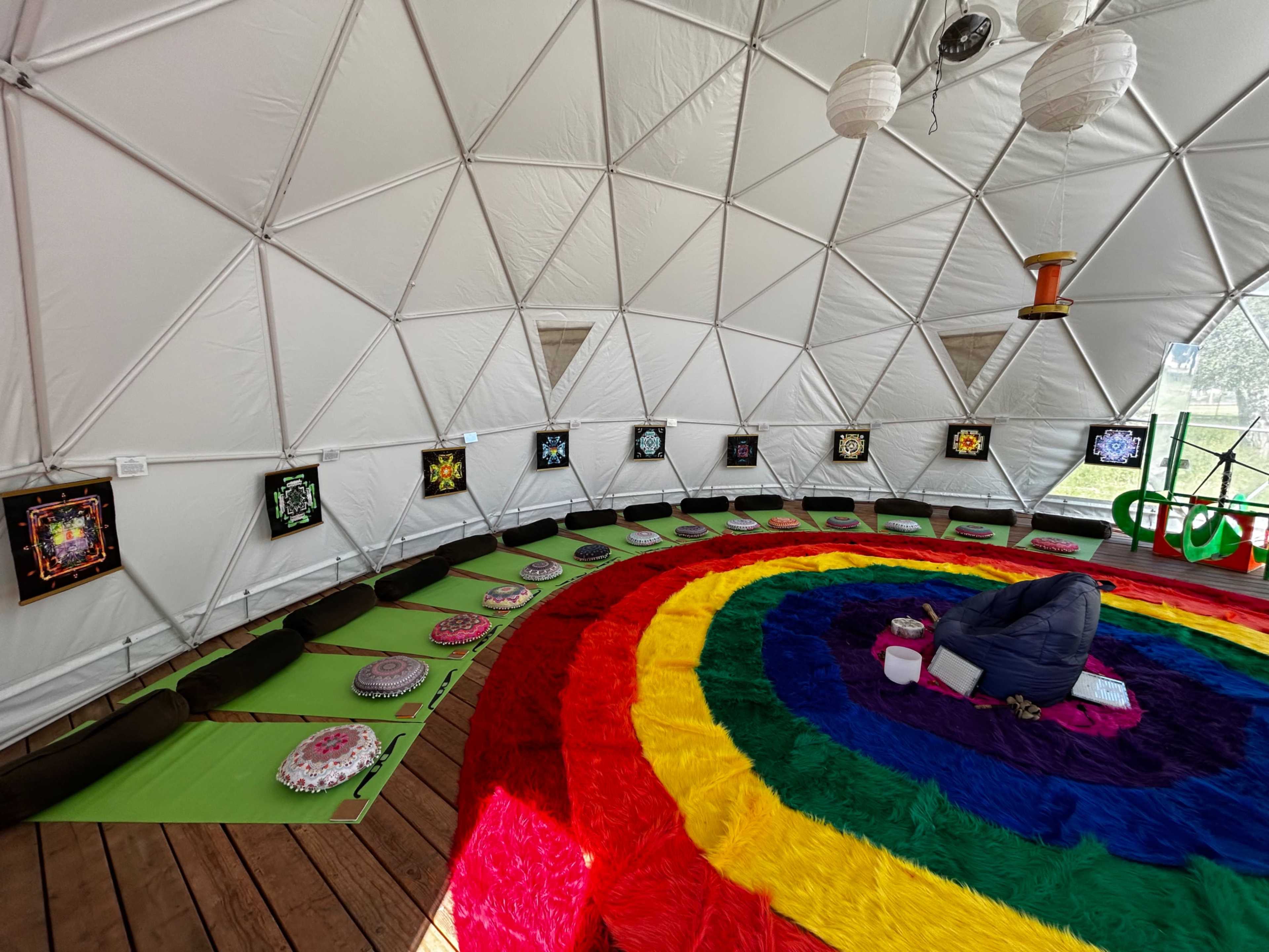 The image depicts the interior of a geodesic dome featuring colorful carpet, wall hangings, and arranged seating on green mats.
