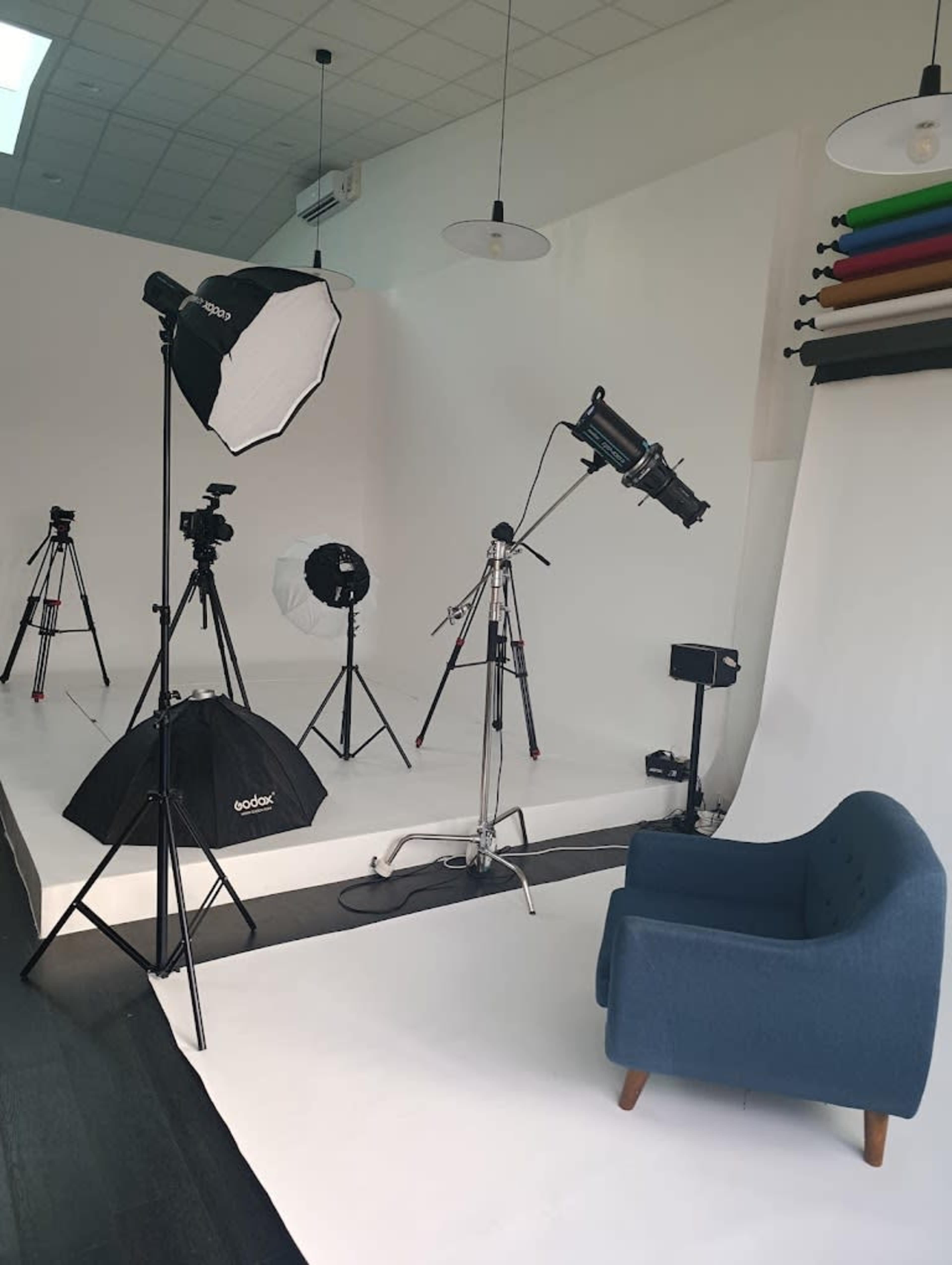 The image shows a photography studio equipped with multiple tripods, lighting setups, and a blue armchair against a plain white backdrop.